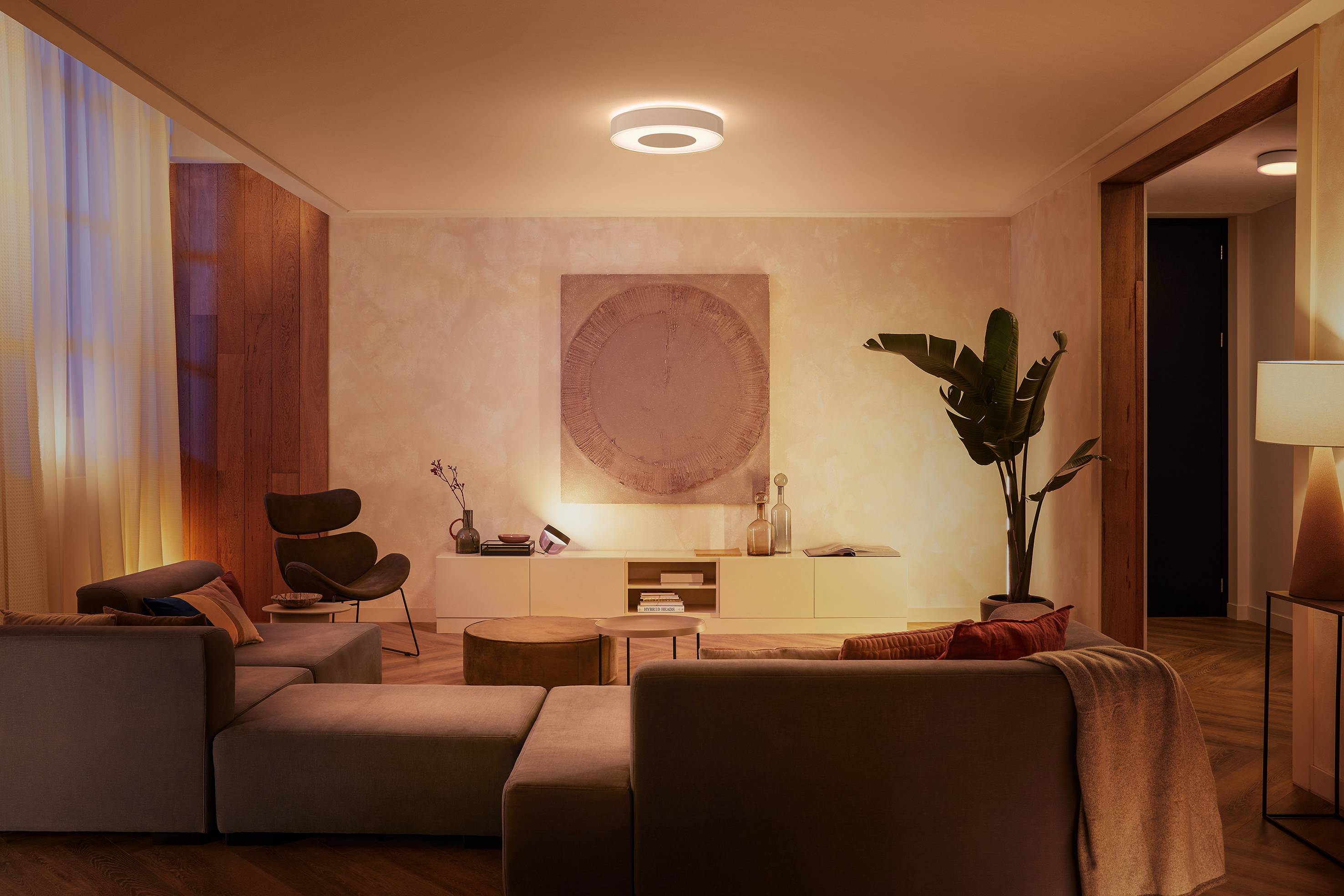 Wohnzimmer bei sanfter Beleuchtung, mit modernen Möbeln, einem großen Sofa, Pflanzen und einem Kunstwerk an der Wand. Gemütliche Atmosphäre.