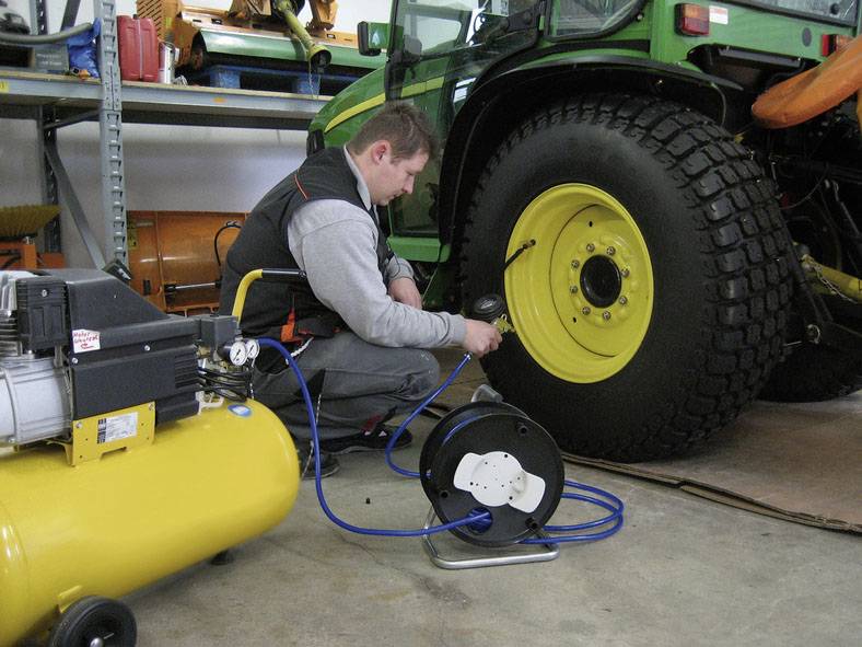 Ein Mechaniker pumpt Luft in einen großen Reifen eines grünen Traktors in einer Werkstatt auf, ein gelber Luftkompressor steht daneben.
