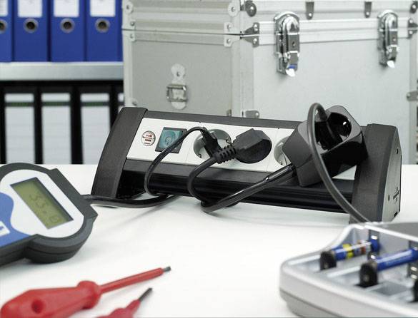 Elektrische Geräte und Messinstrumente auf einem Tisch, umgeben von Aktenordnern und einem silbernen Koffer im Hintergrund.