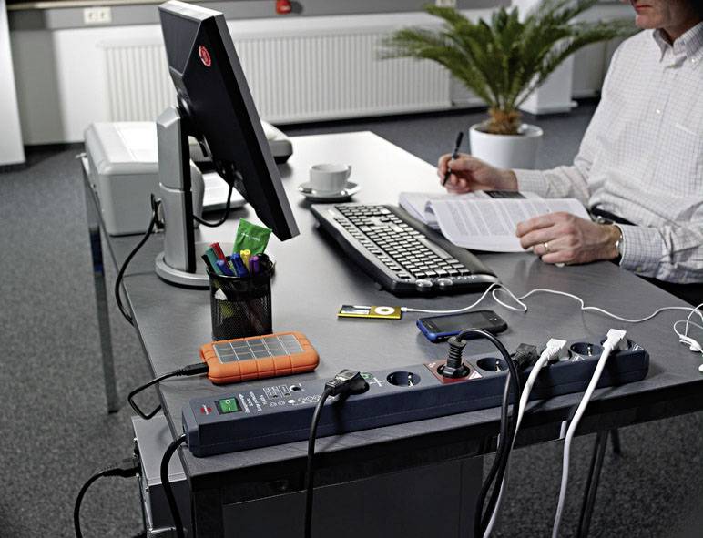 Eine Person arbeitet an einem Schreibtisch mit Computer, Dokumenten und Büromaterial. Mehrere Geräte sind an eine Steckdosenleiste angeschlossen.