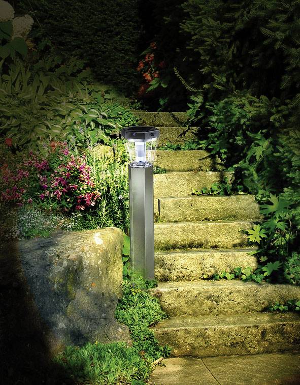 Steinweg im Garten mit grünen Pflanzen und Blumen, beleuchtet von einer Solarlaterne auf einem Pfahl.