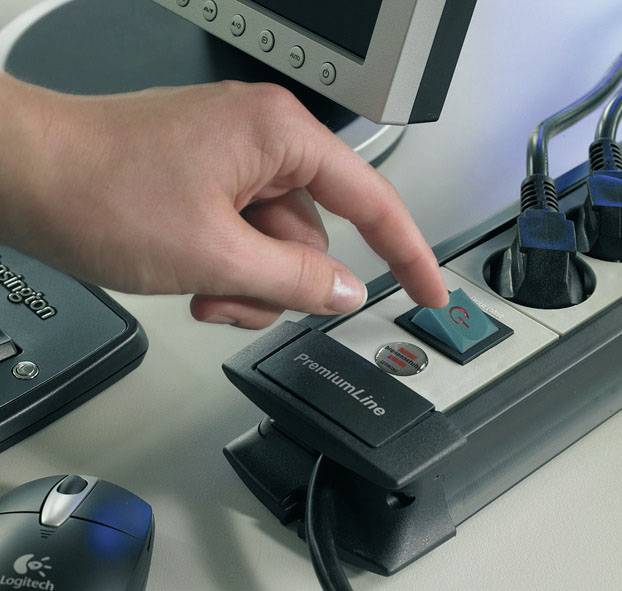 Eine Hand drückt den roten Knopf einer Steckdosenleiste neben einem Computerbildschirm. Mehrere Kabel sind angeschlossen.