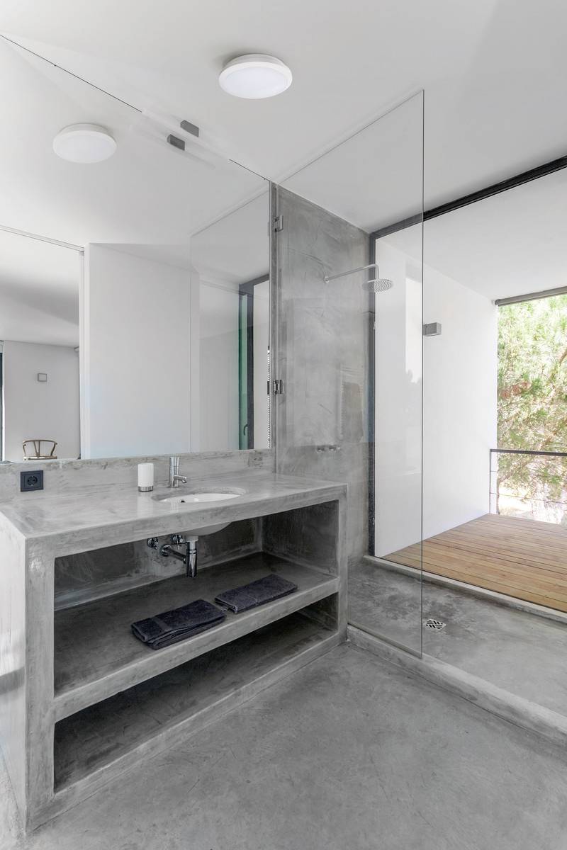 Badezimmer mit modernen Betonelementen, Waschbecken, Dusche mit Glasabtrennung und Blick auf einen Balkon mit Holzboden.