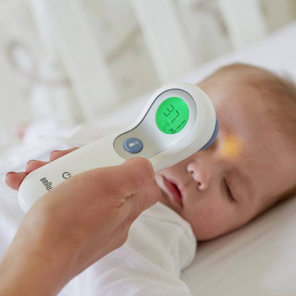 Ein Baby liegt auf einem Bett, während eine Person ein digitales Infrarot-Thermometer an seiner Stirn hält und seine Temperatur misst.