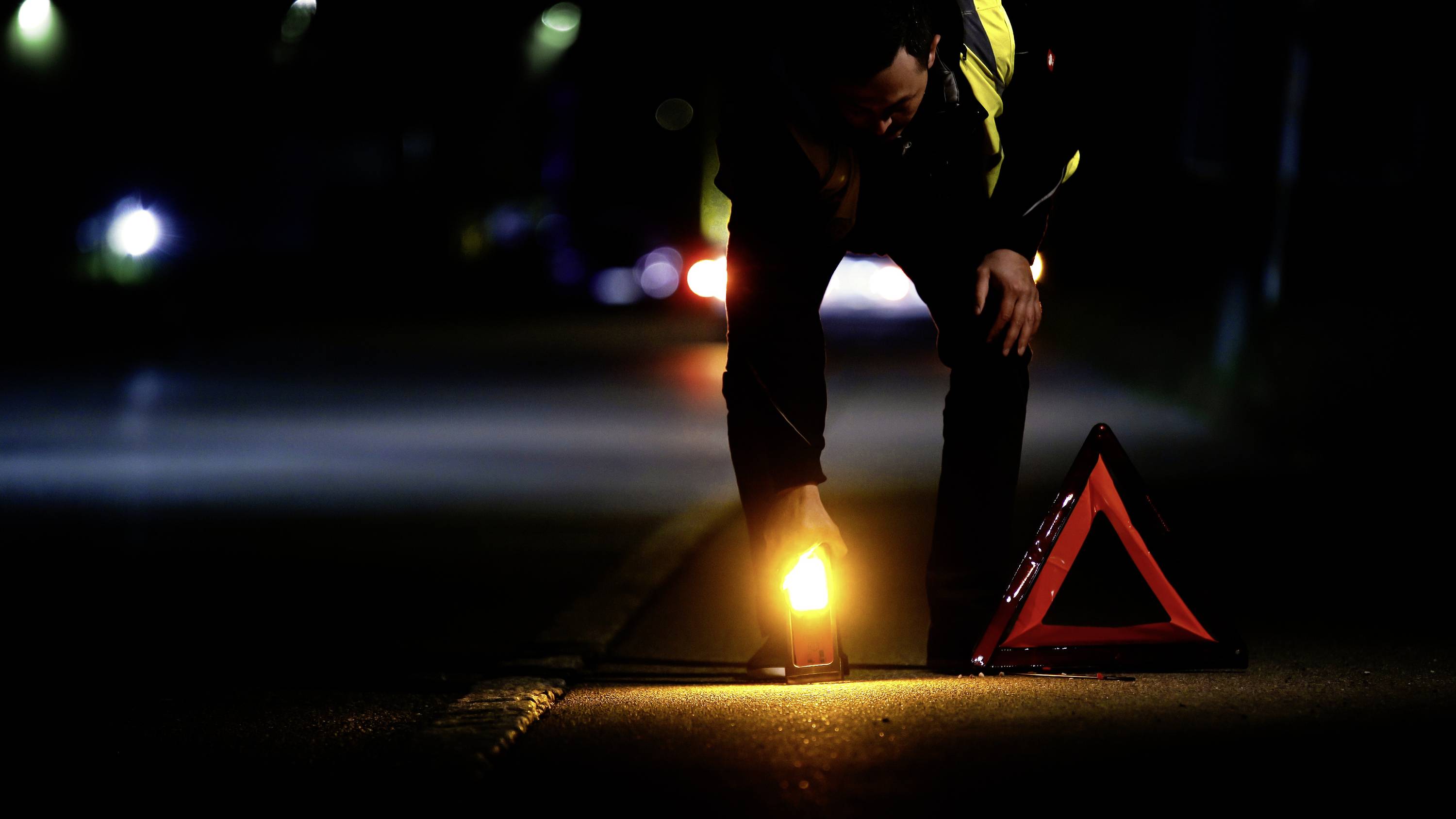 Eine Person platziert nachts ein leuchtendes Warnsignal und ein Warndreieck auf der Straße, um vor einem potenziellen Unfall zu warnen.