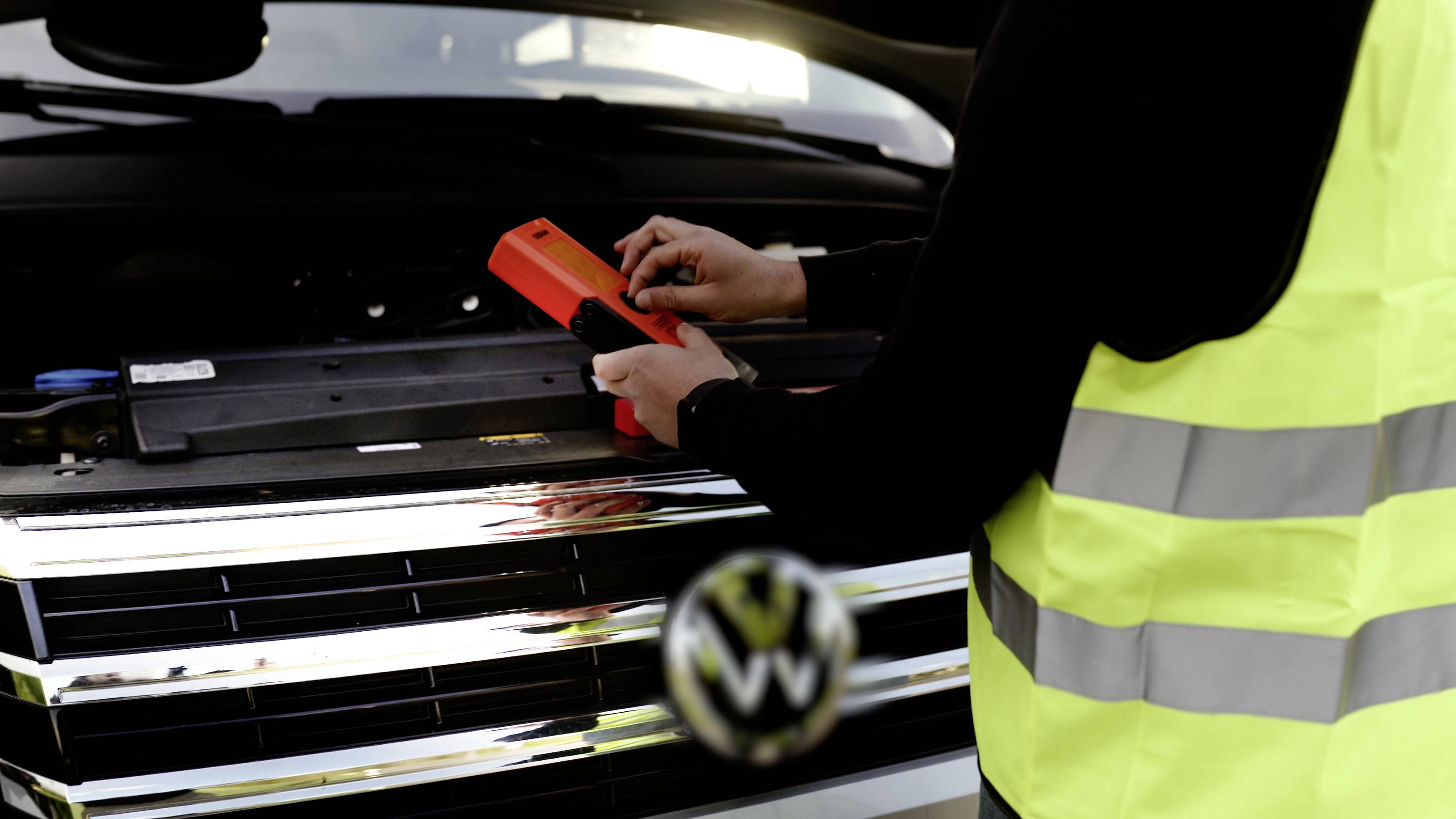 Eine Person trägt eine Warnweste, öffnet die Motorhaube eines Autos und prüft mit einem Messgerät den Motorraum.