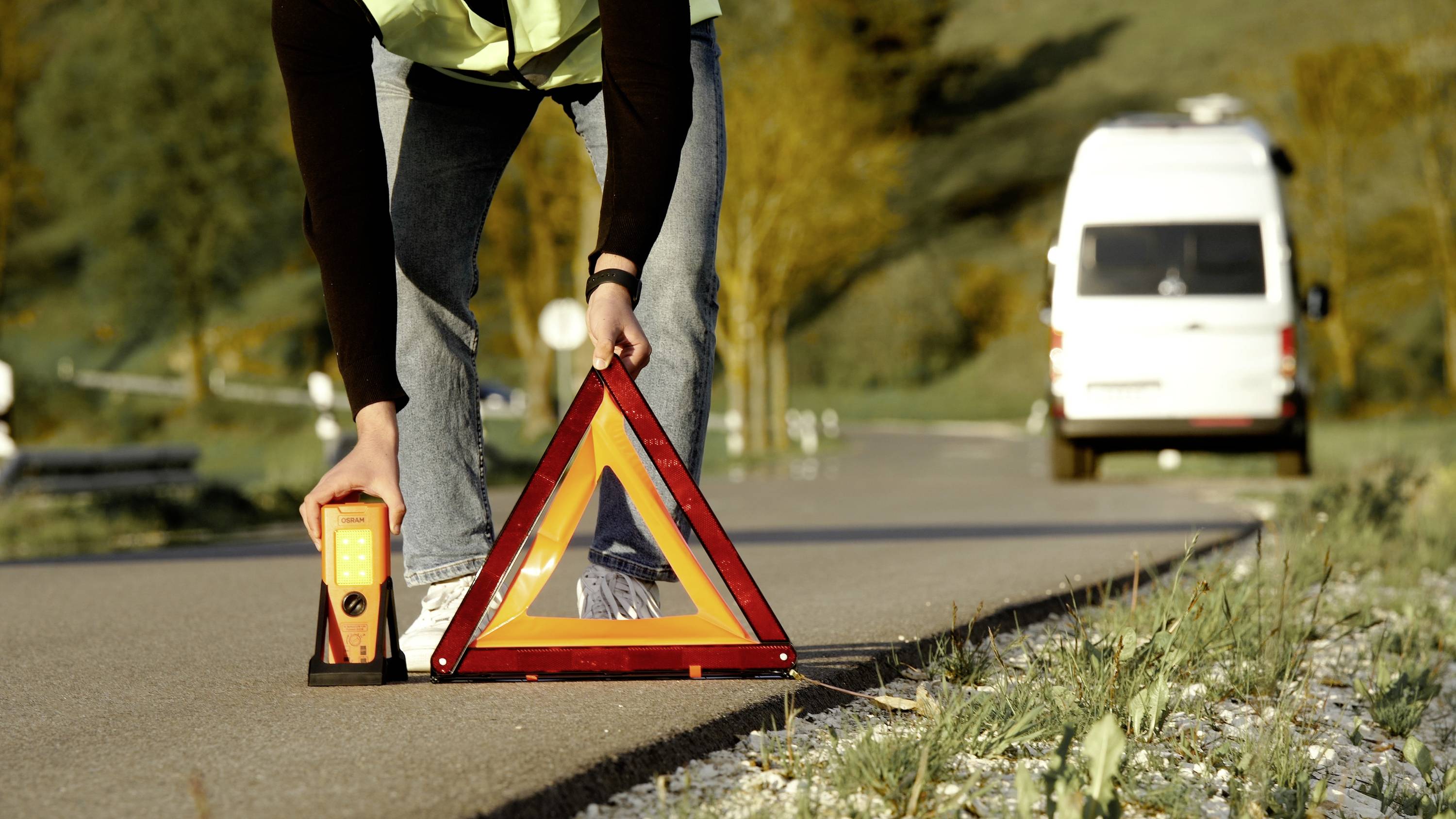 Person stellt ein Pannendreieck auf der Straße auf; im Hintergrund ein geparktes Fahrzeug. Szenario einer Autopanne oder eines Unfalls.