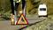 Person stellt ein Pannendreieck auf der Straße auf; im Hintergrund ein geparktes Fahrzeug. Szenario einer Autopanne oder eines Unfalls.