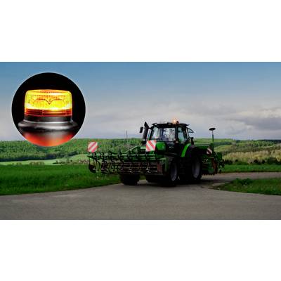 Ein Traktor auf einer Straße auf einem Feld mit einer Nahaufnahme eines gelben Warnlichts. Wälder und Himmel im Hintergrund.