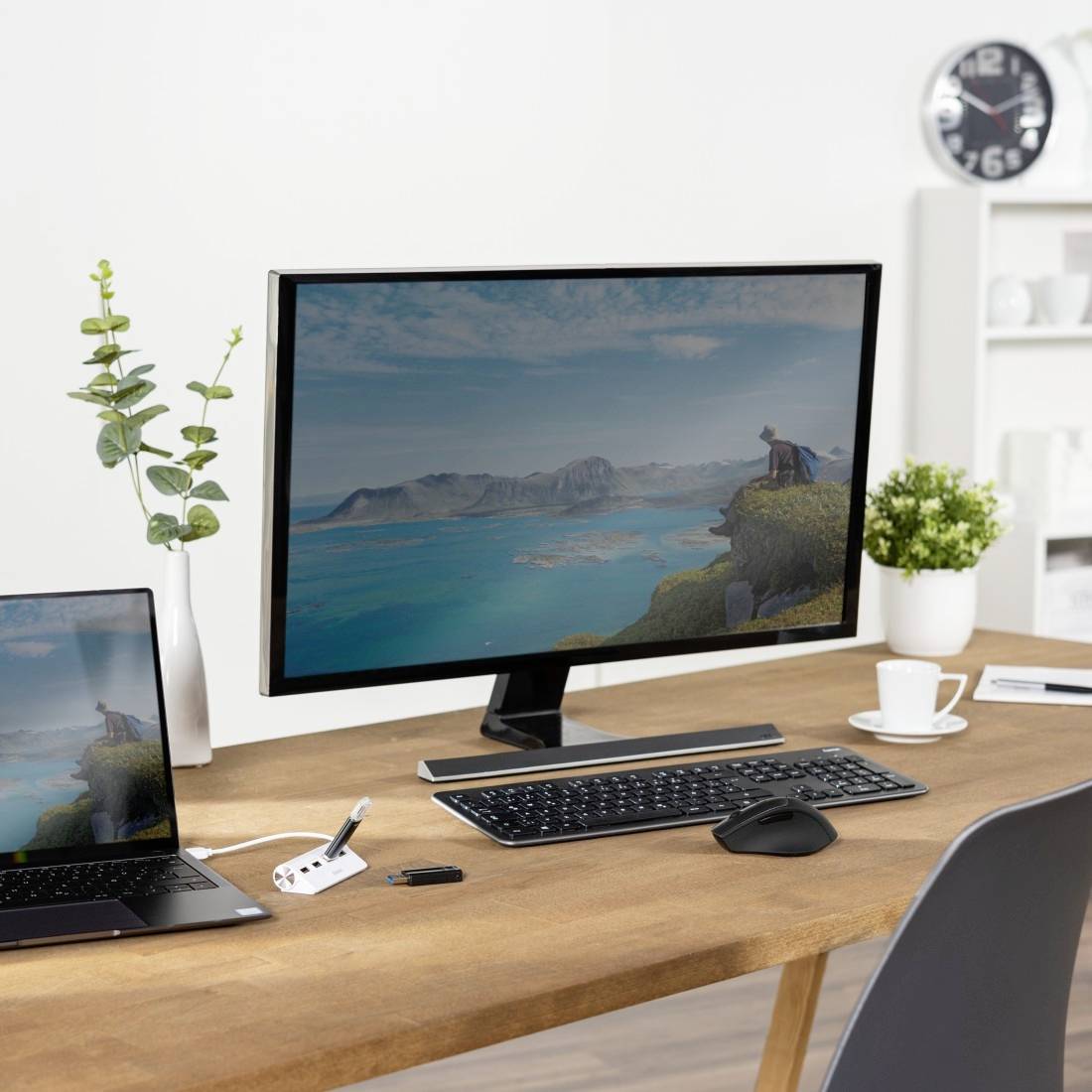 Ein Schreibtisch mit einem Computerbildschirm, der eine Landschaft zeigt, umgeben von Pflanzen, Laptop, Tastatur, Maus und Kaffeetasse.