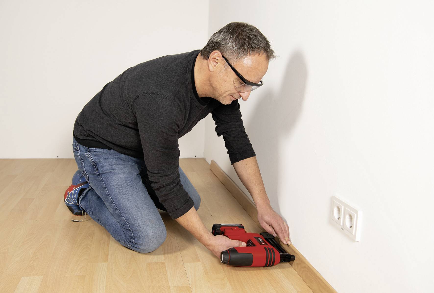 Ein Mann kniet auf Laminatboden und benutzt ein rotes Elektrowerkzeug, um Fußleisten an einer weißen Wand zu befestigen.