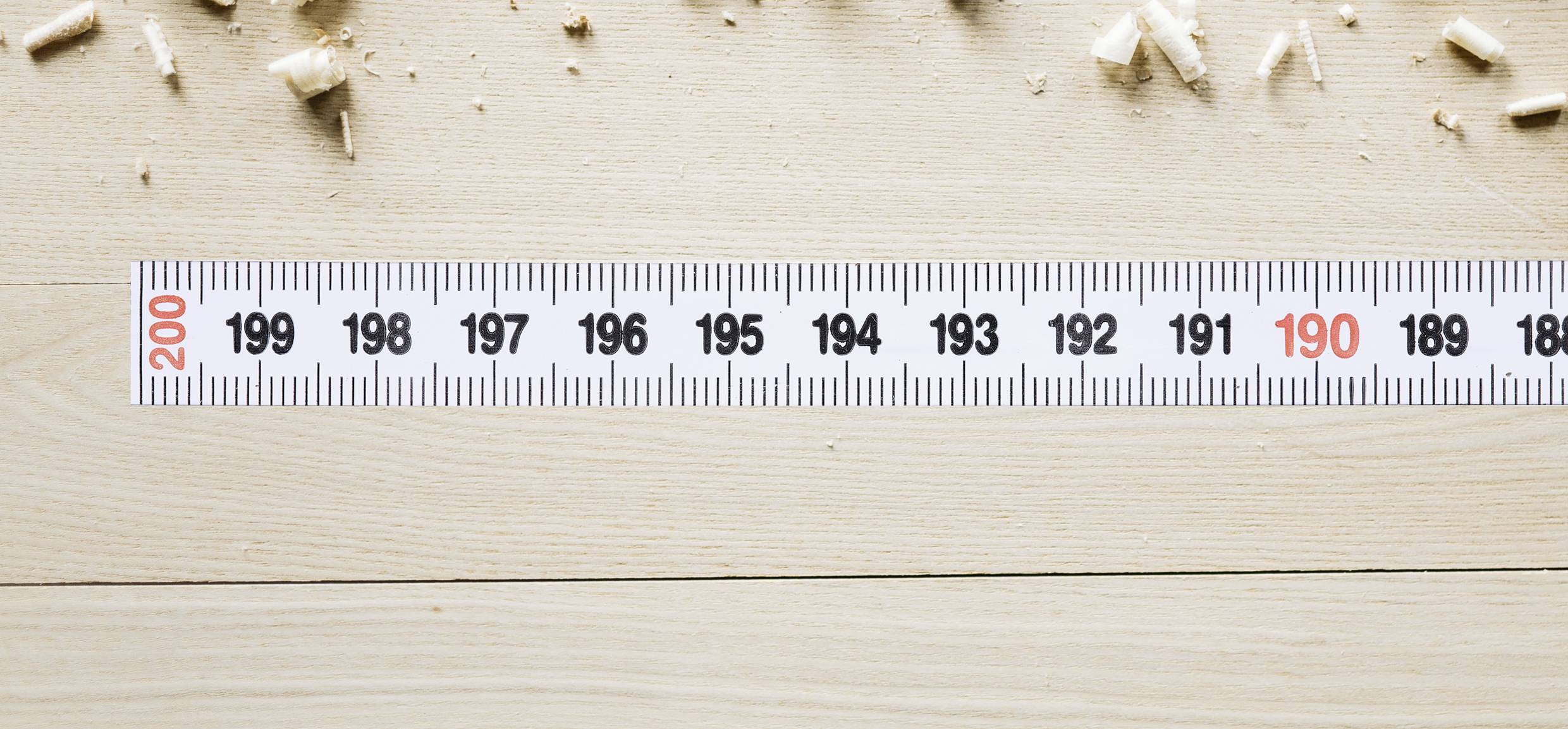 Ein Maßband liegt auf einem Holztisch, zeigt 190 cm in roter Schrift an, umgeben von Holzspänen am oberen Rand.