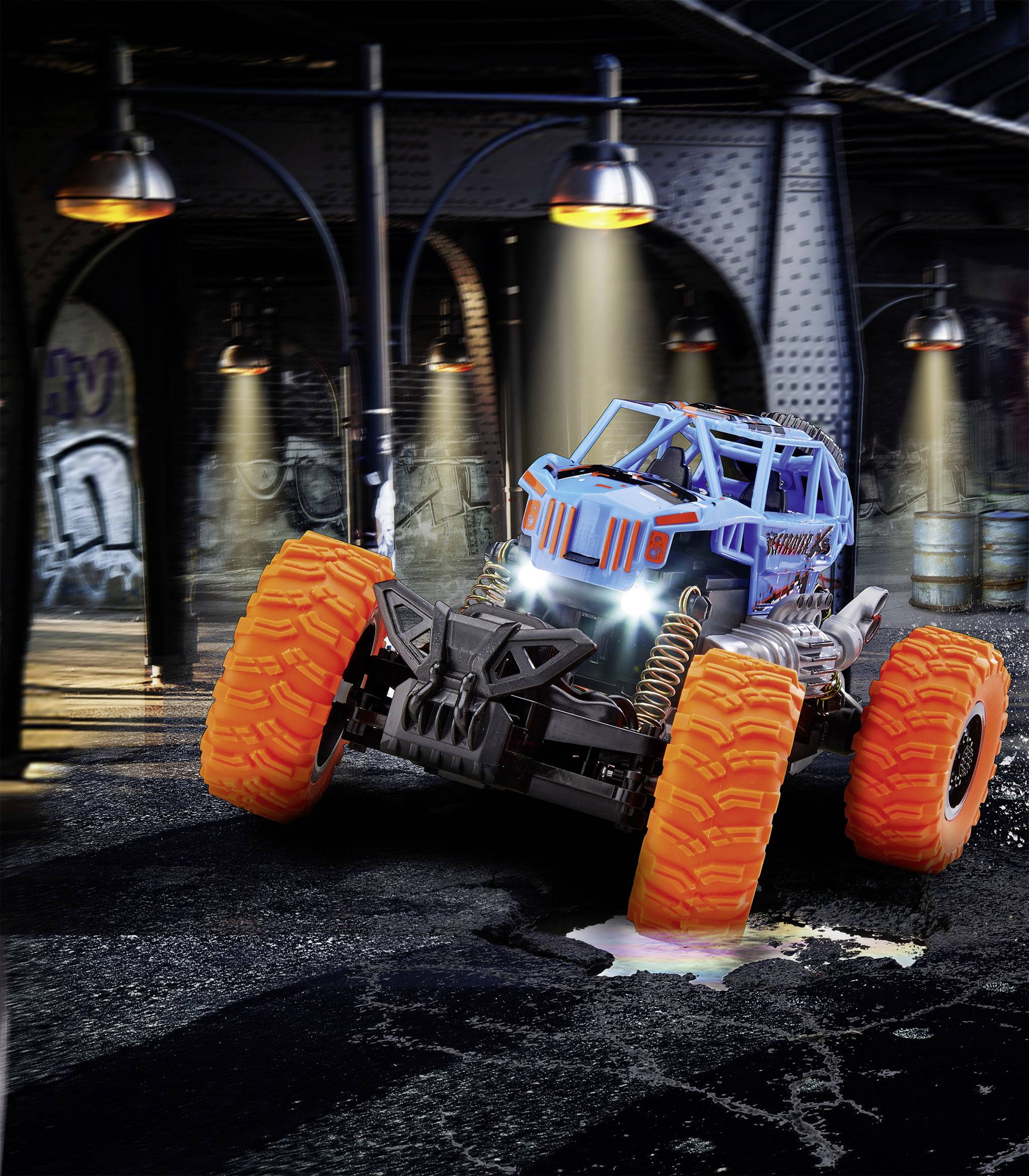 Geländewagen-Spielzeugauto mit großen orangefarbenen Reifen fährt über nasse Betonfläche in industrieller Umgebung, nachts beleuchtet.