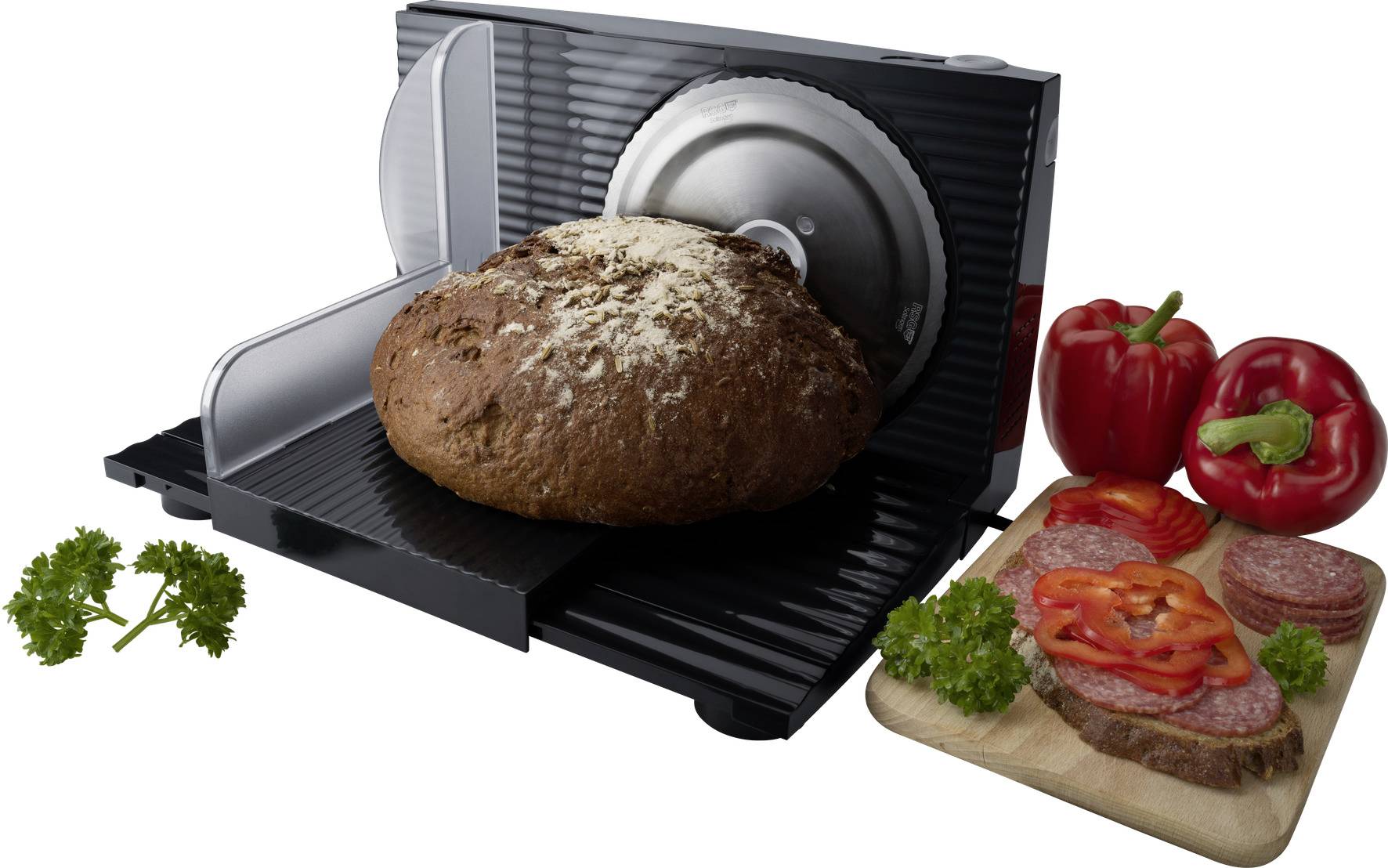Eine elektrische Brotschneidemaschine schneidet dunkles Brot. Daneben liegen rote Paprika, Salami und Petersilie auf einem Holzbrett.