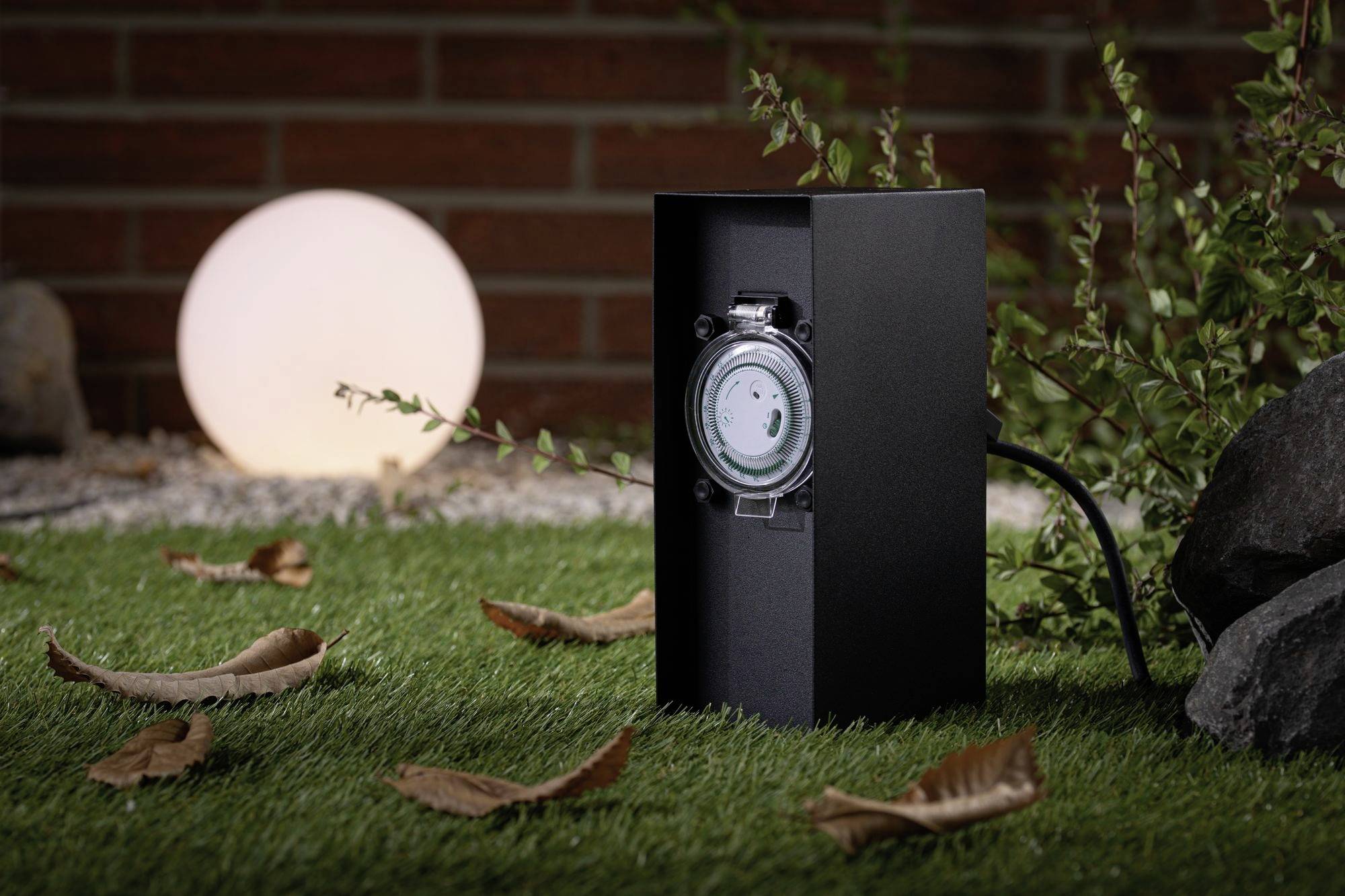 Eine schwarze Gartensteckdose mit Timer auf Rasen, umgeben von Blättern und Pflanzen; im Hintergrund ein leuchtender Kugellampe.