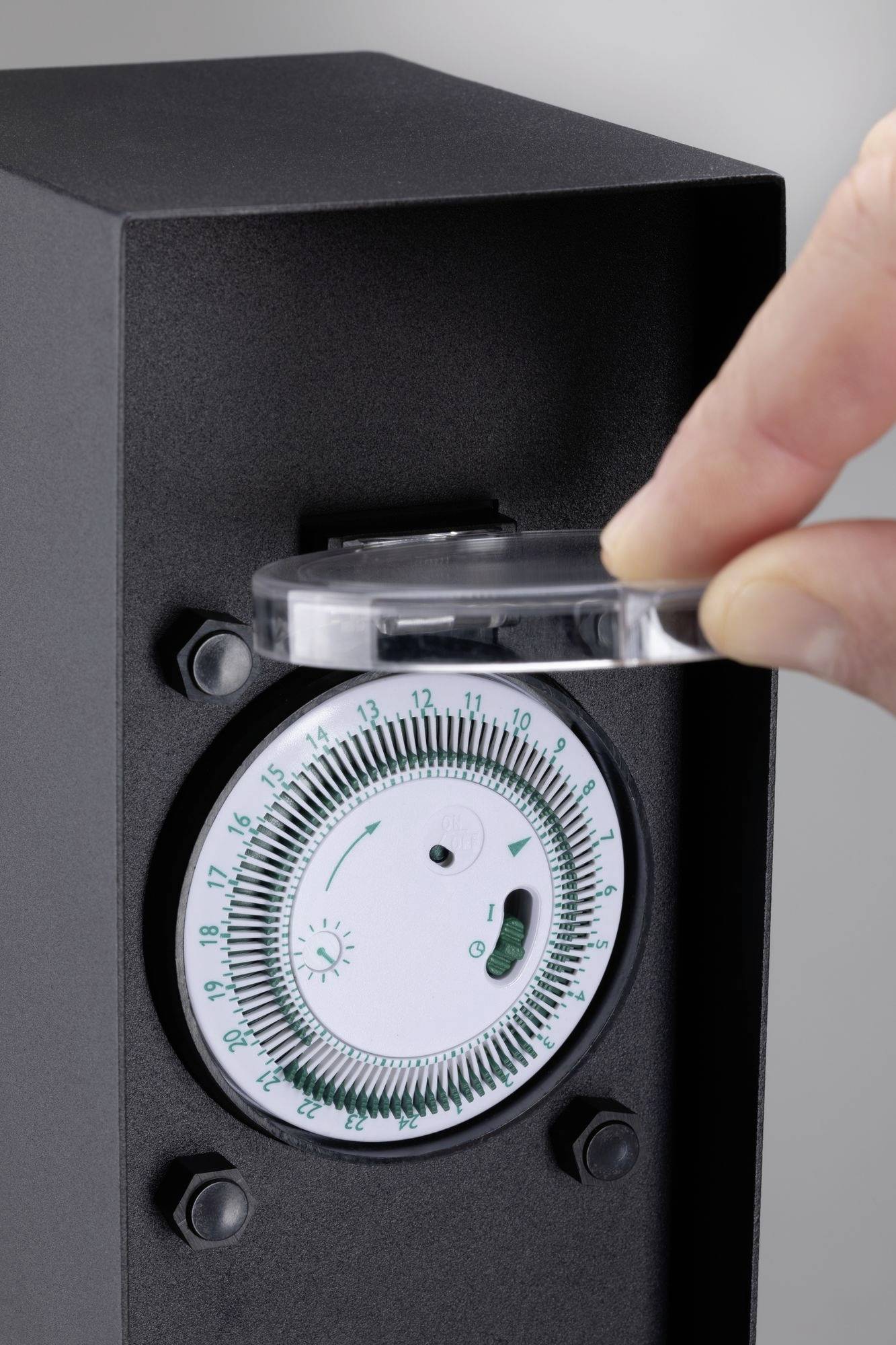 Eine Hand dreht die Abdeckung eines schwarzen Uhrenmechanismus, auf dem ein Zifferblatt mit Zahlen und Markierungen zu sehen ist.