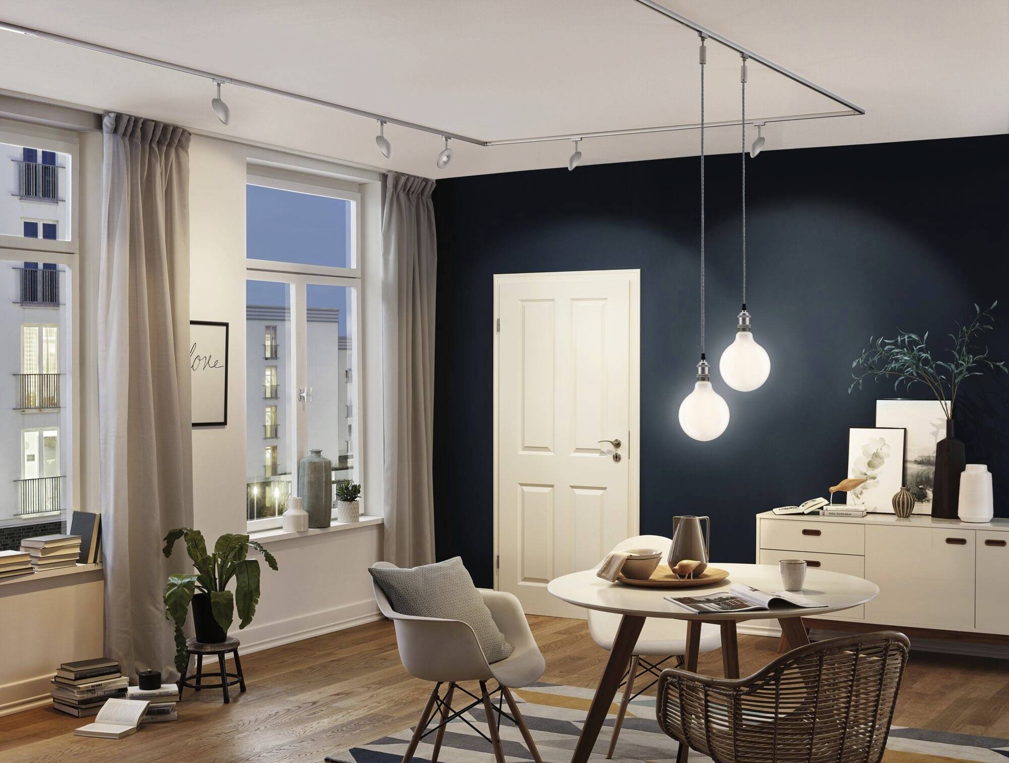 Ein gemütliches Wohnzimmer mit Holzboden, einem runden Tisch und Hängelampen. Fenster zeigen städtische Gebäude bei Dämmerung.