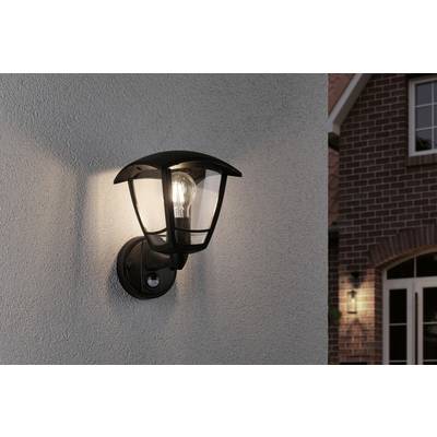 Außenlampe an einer Hauswand, die in der Dämmerung eingeschaltet ist. Im Hintergrund ein Ziegelhaus mit erleuchtetem Fenster.