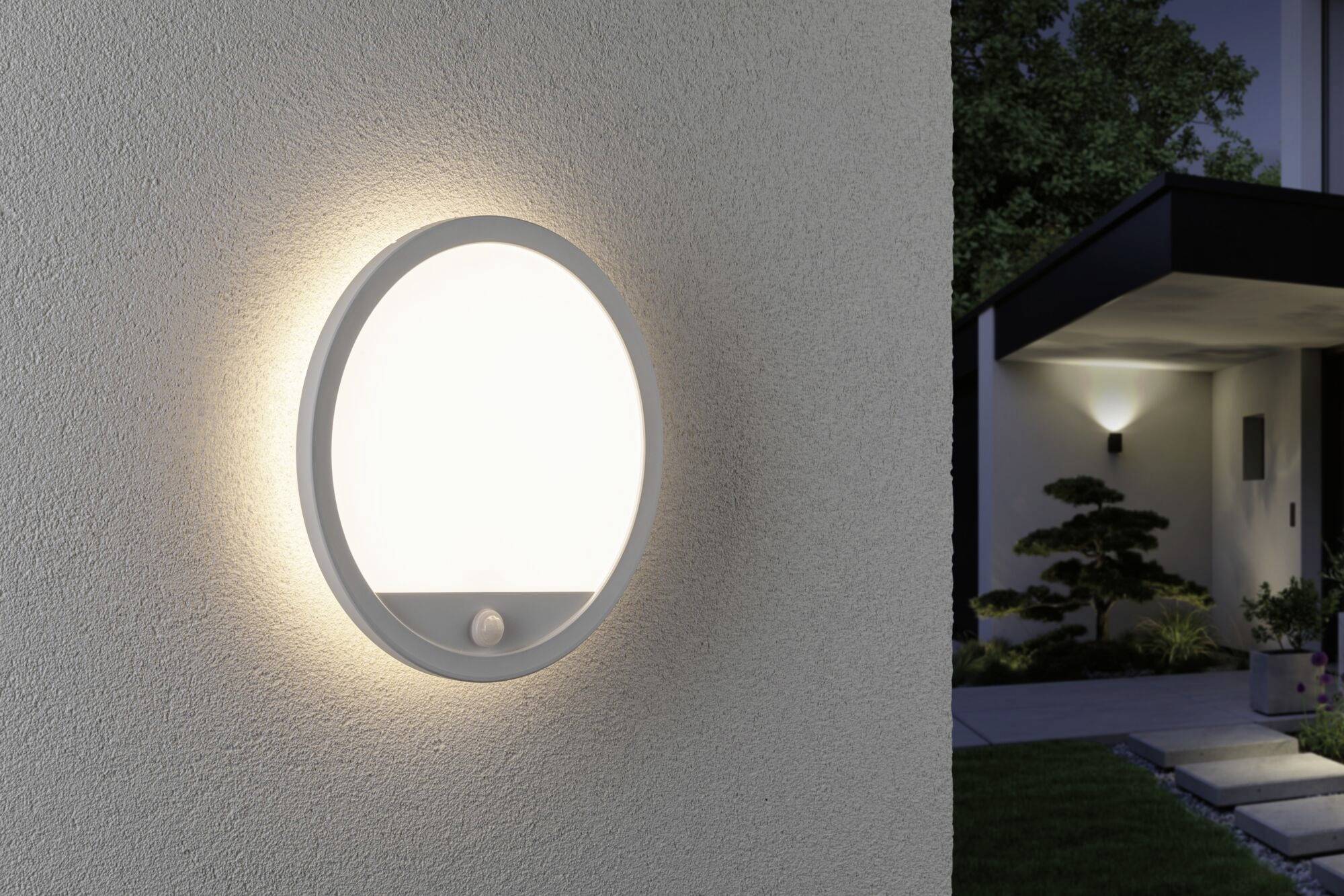 Eine moderne runde Außenlampe mit Bewegungsmelder leuchtet an einer Hauswand. Im Hintergrund ein Eingang mit beleuchtetem Weg.