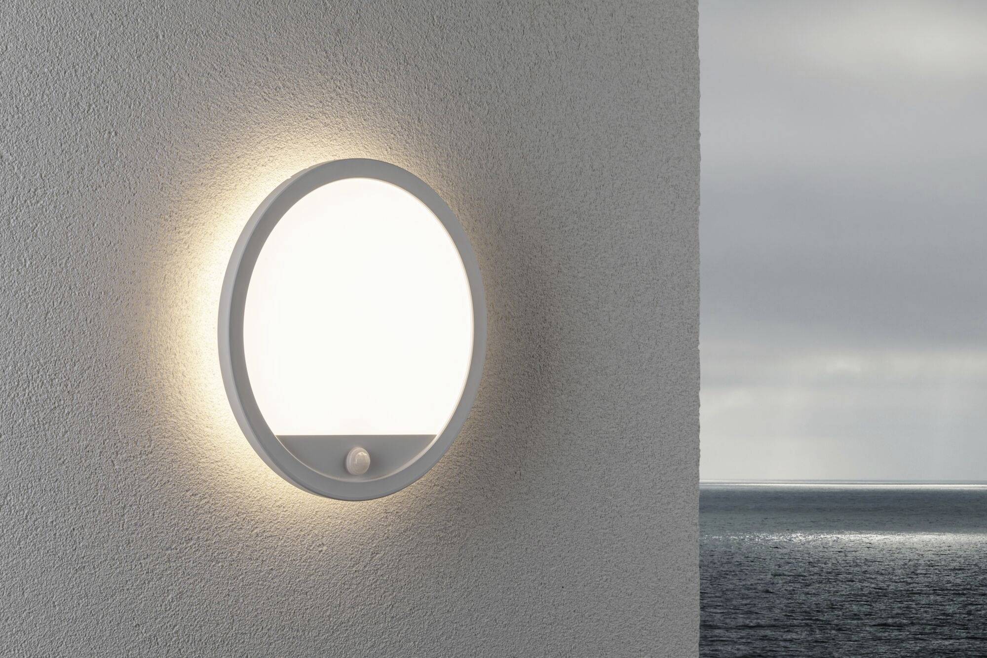Eine runde Wandleuchte leuchtet an einer weißen Wand. Draußen ist der Himmel bewölkt über dem Meer.