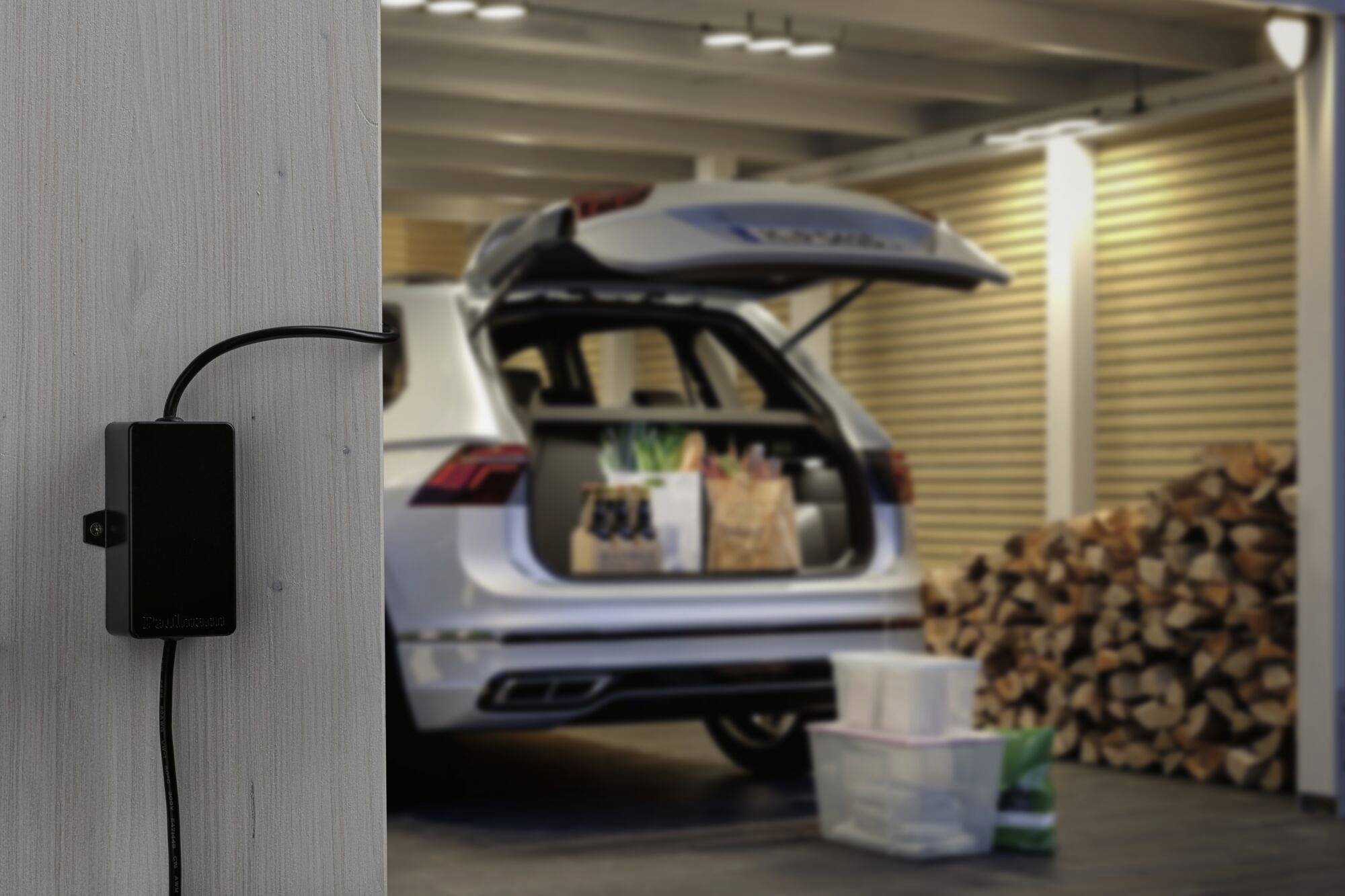 Ein geparktes Auto im Carport mit geöffneter Heckklappe. Im Kofferraum stehen Einkaufstüten. Im Vordergrund ist ein Ladegerät an der Wand.