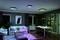 Wohnzimmer mit moderner Einrichtung; beleuchtet von bunten LED-Deckenlichtern. Heller Teppich, Sofa, Esstisch und Pflanzen als Dekor.