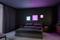 Ein modernes Schlafzimmer mit einem großen Bett, dunklen Wänden und Vorhängen. An der Wand hängen drei Leuchtbilder in Blau und Rosa.