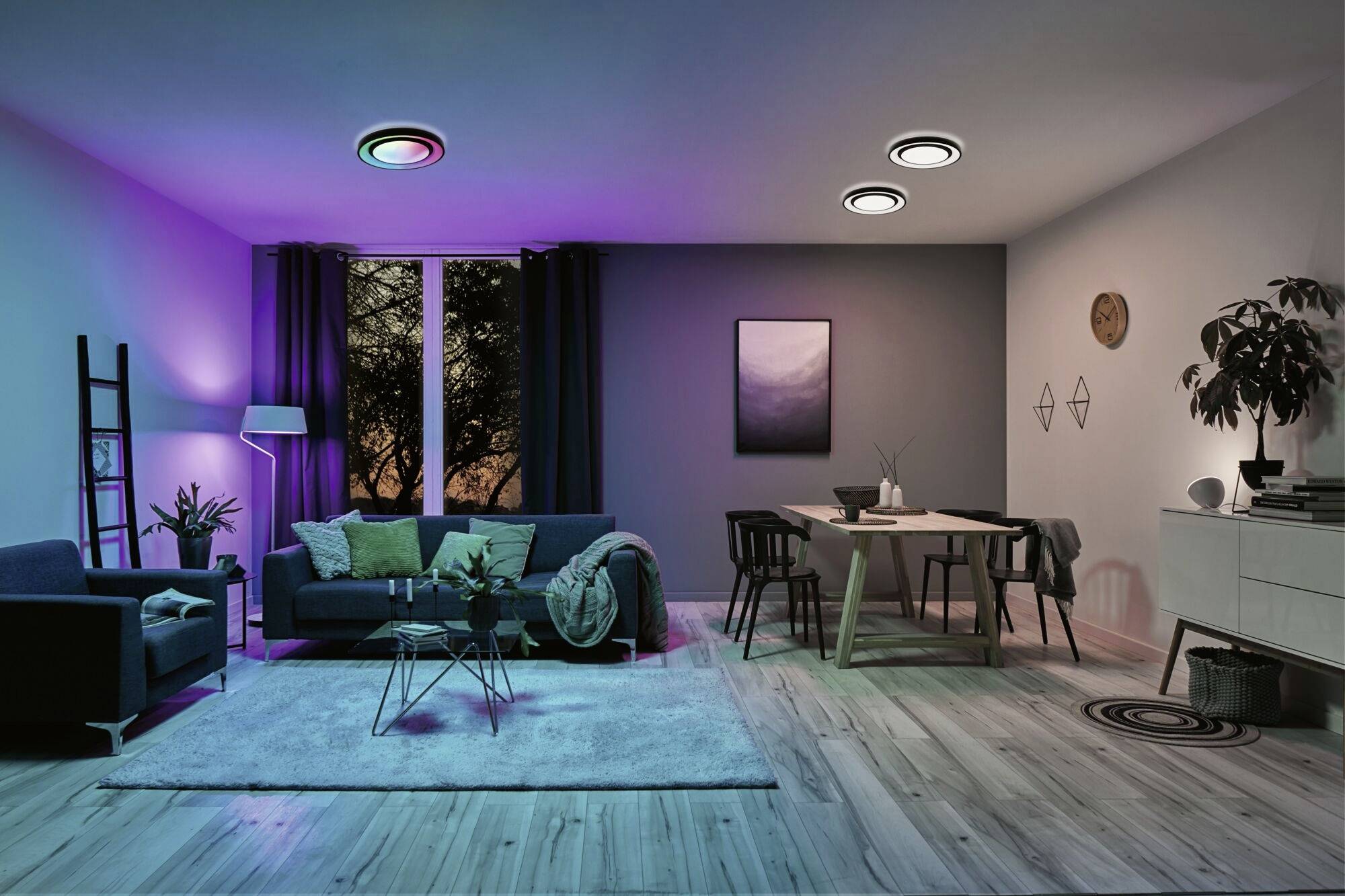 Modernes Wohnzimmer mit farbigem Stimmungslicht. Ein Sofa und Tisch gruppiert, Fenster mit Vorhang zeigt Abendhimmel.