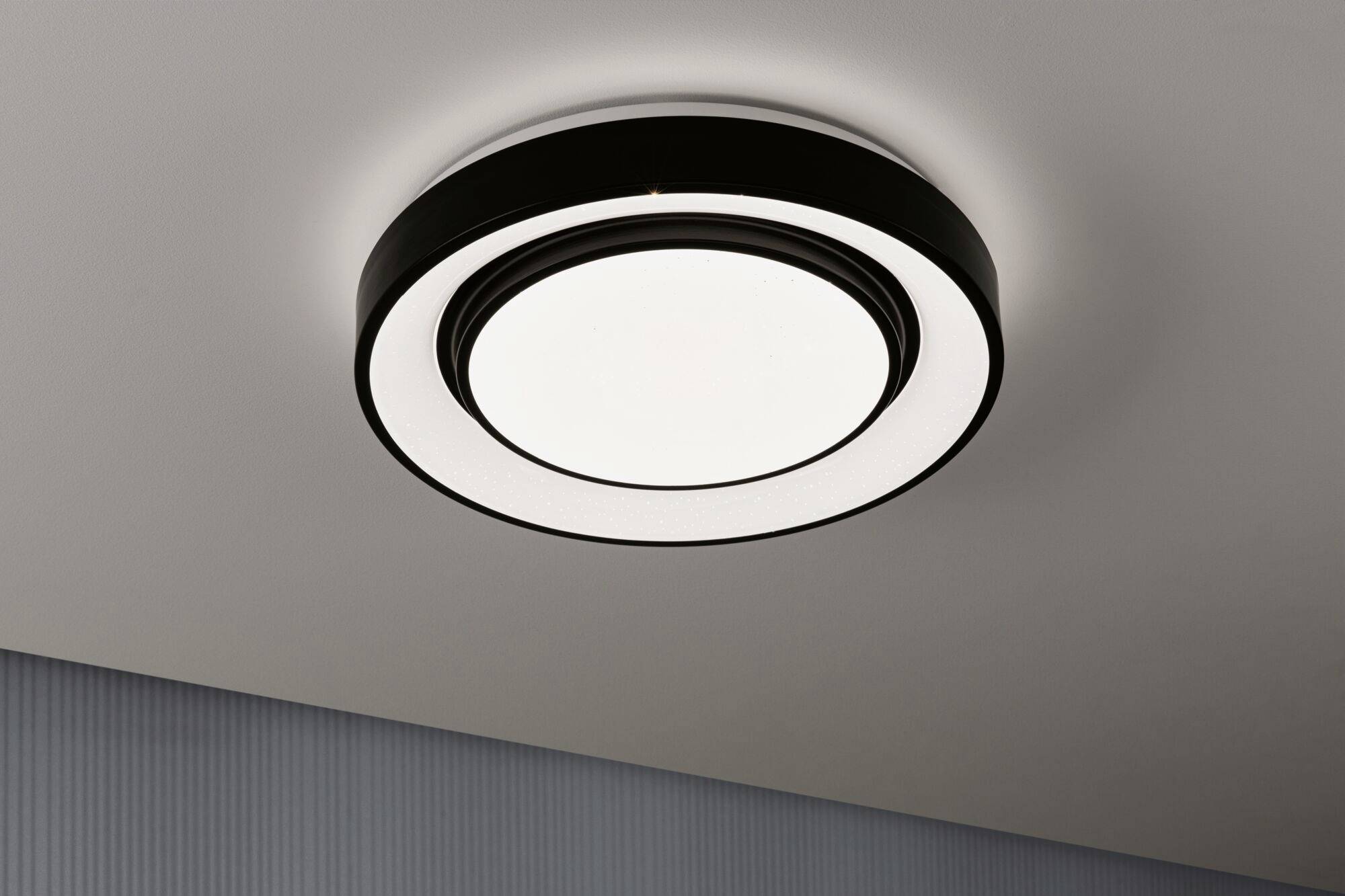 Runde, moderne Deckenlampe mit schwarzem Rahmen und hellem Licht, an einer weißen Decke montiert.