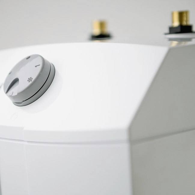 Ein weißes Heizgerät mit einem Thermostatknopf und zwei Anschlussventilen auf der Oberseite. Gerät steht vor einem neutralen Hintergrund.