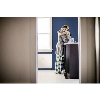 Eine Person in Schlafanzug umarmt eine andere in der Küche. Sie stehen nahe der Spüle. Hintergrund ist eine blaue Wand und ein Fenster.