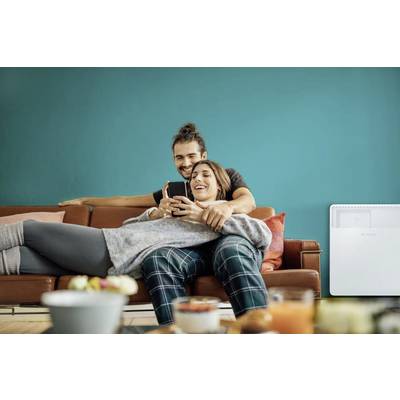 Ein lächelndes Paar sitzt auf einem Sofa, eng umschlungen. Beide schauen auf ein Smartphone, umgeben von gemütlicher Wohnzimmeratmosphäre.