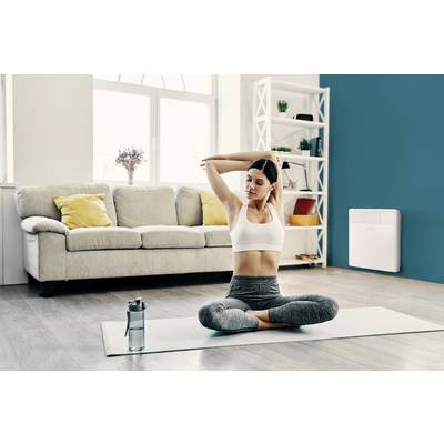 Frau in Sportkleidung dehnt sich auf Yogamatte in einem modernen Wohnzimmer. Hintergrund zeigt Sofa, Kissen und Regal.