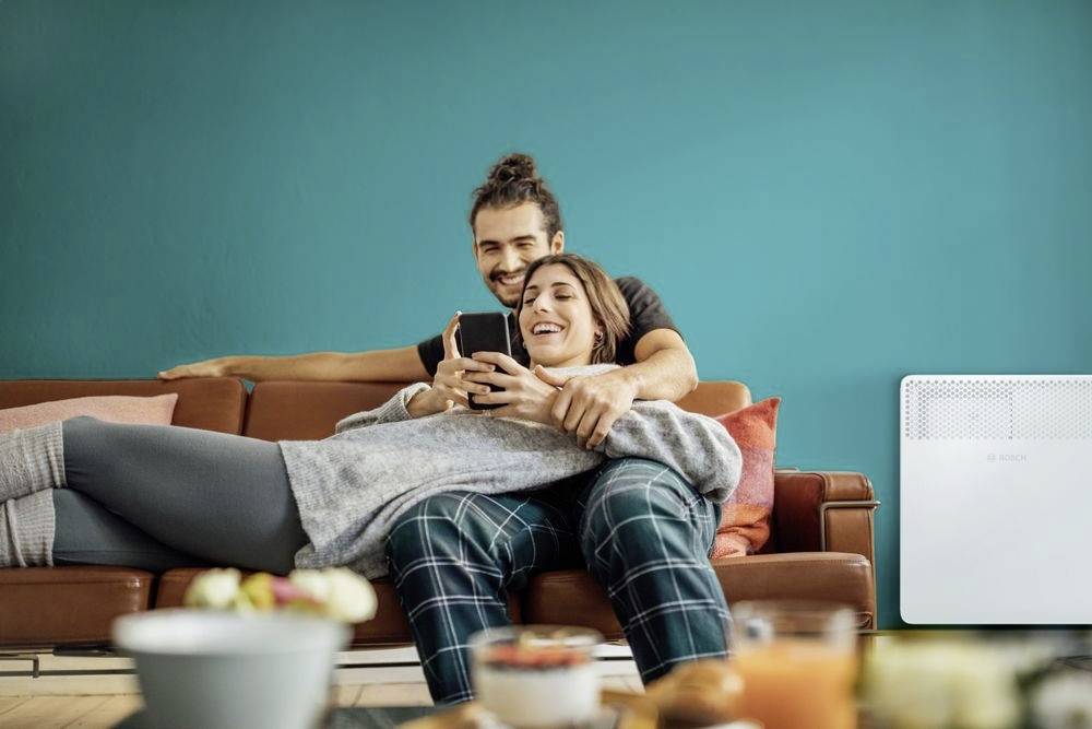 Ein Paar sitzt lächelnd auf einem Sofa. Die Frau hält ein Smartphone, während der Mann sie von hinten umarmt. Wohnzimmeratmosphäre.