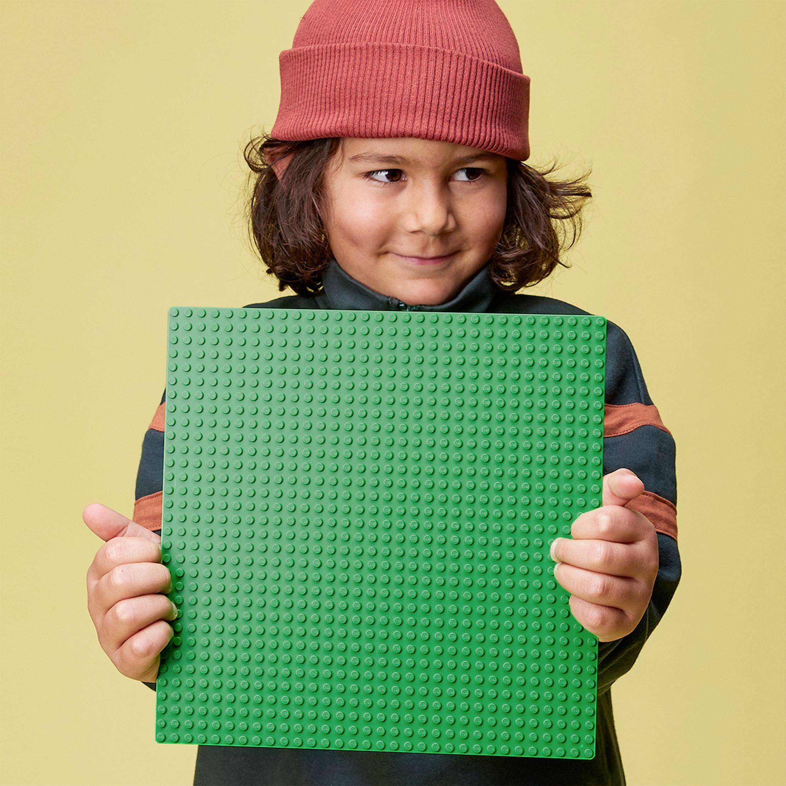 Ein Kind mit roter Mütze hält eine grüne Lego-Grundplatte vor einem gelben Hintergrund. Es lächelt leicht und schaut zur Seite.