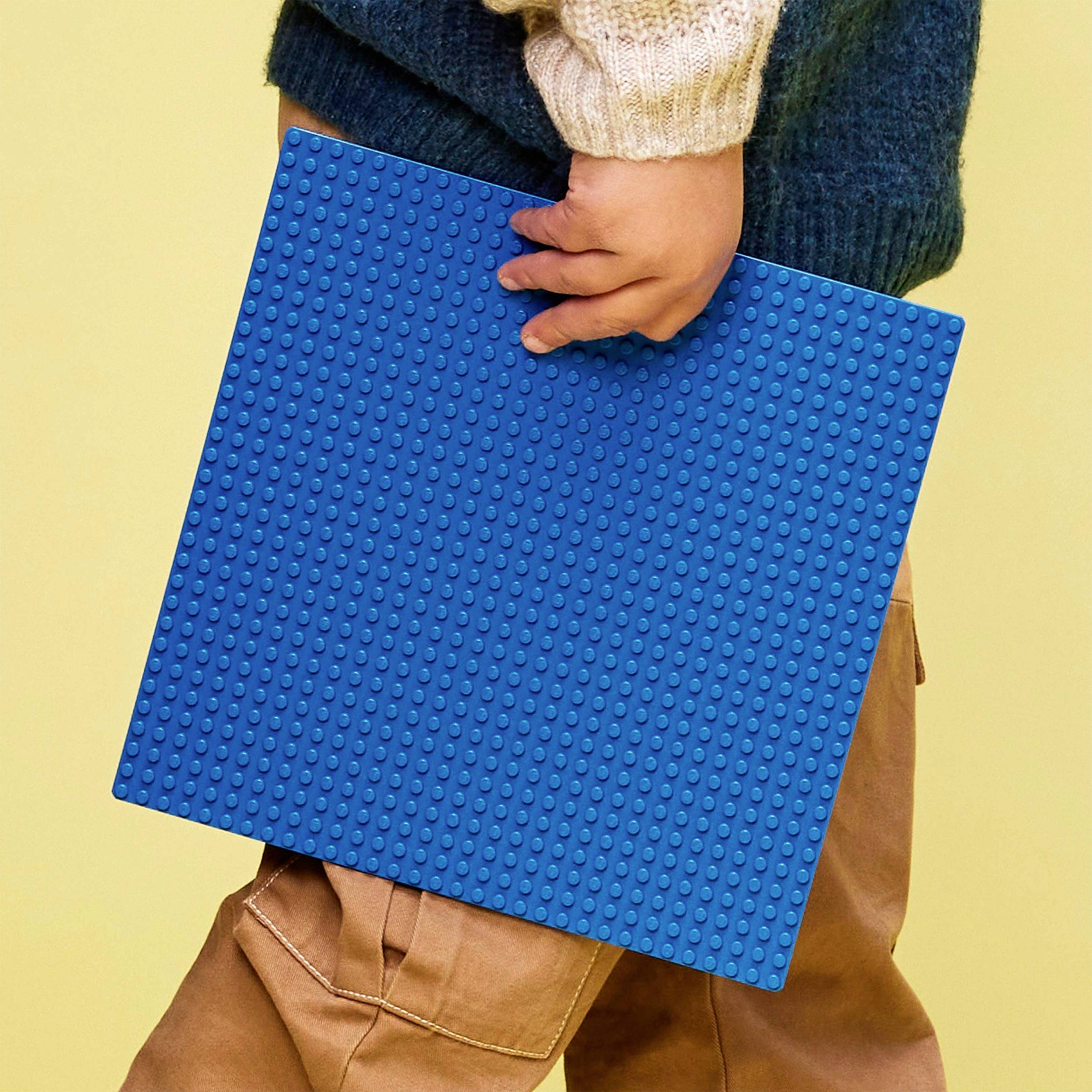 Ein Kind hält eine große blaue Bauplatte aus Kunststoff. Es trägt eine braune Hose und einen blauen Pullover mit beigem Ärmel.