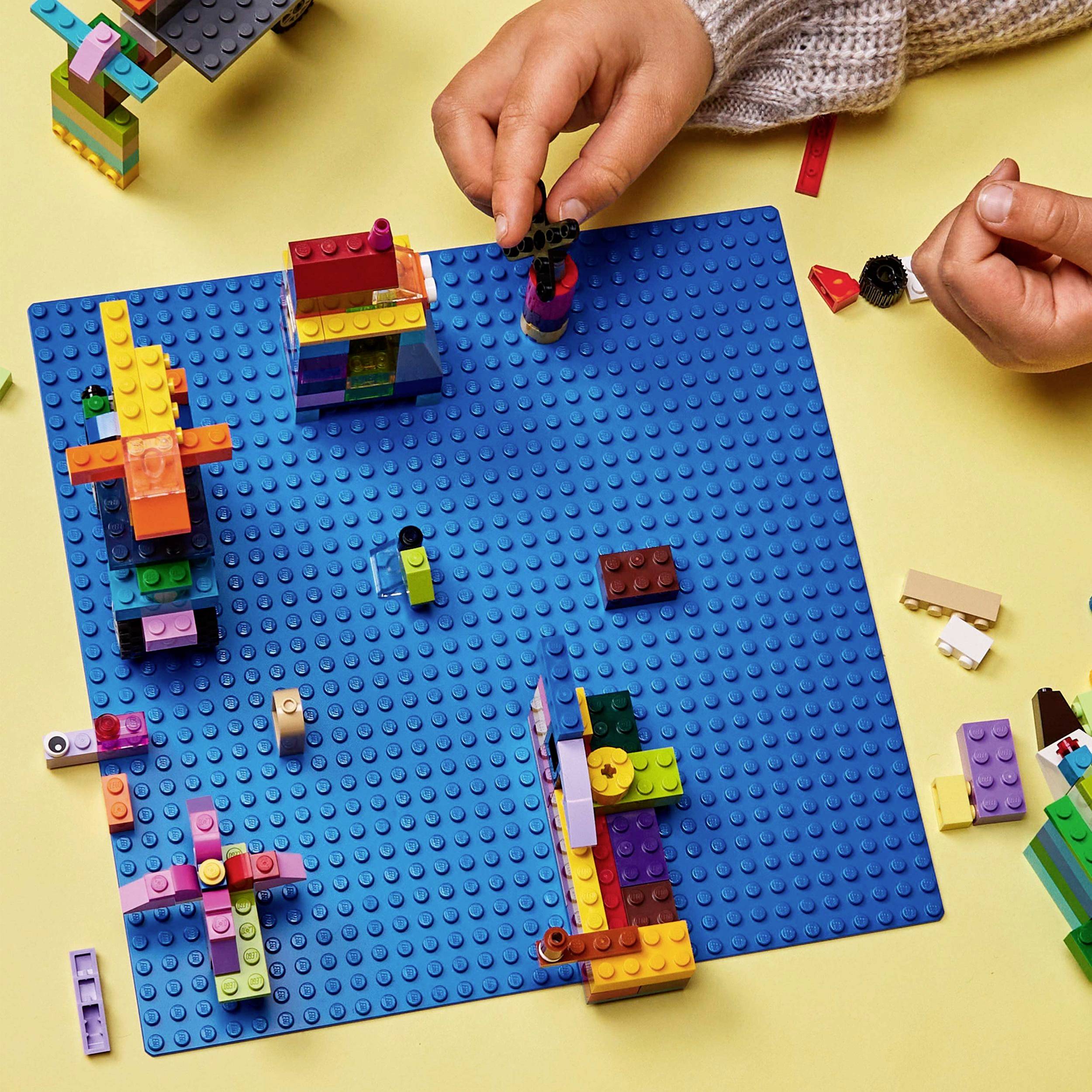 Eine Person baut ein buntes Lego-Gebilde auf einer blauen Grundplatte auf einem Tisch. Mehrere bunte Lego-Teile sind verteilt.