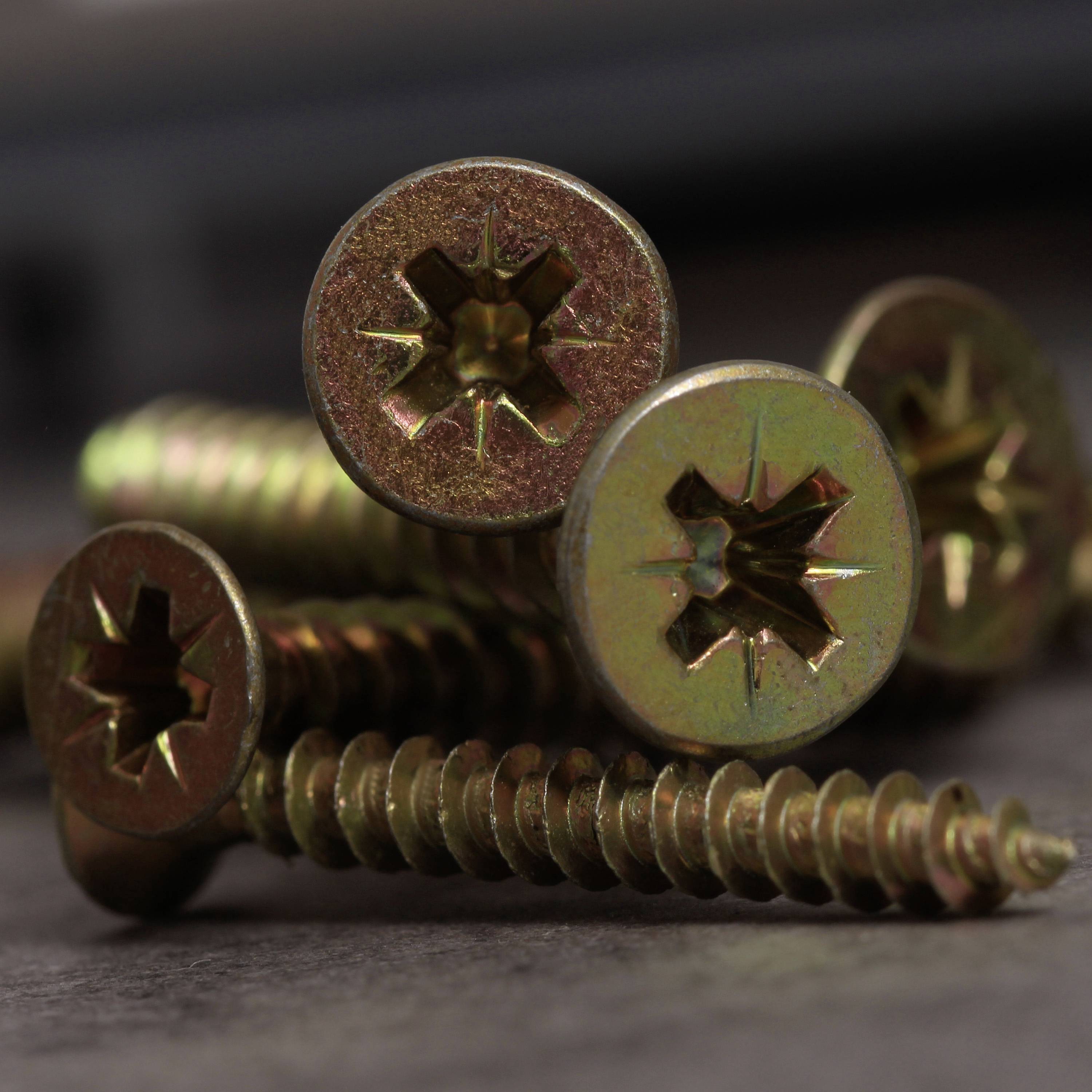 Vier Metallschrauben mit Kreuzschlitzköpfen liegen auf einer dunklen Oberfläche. Sie sind rostfrei und in einem schimmernden Goldton.