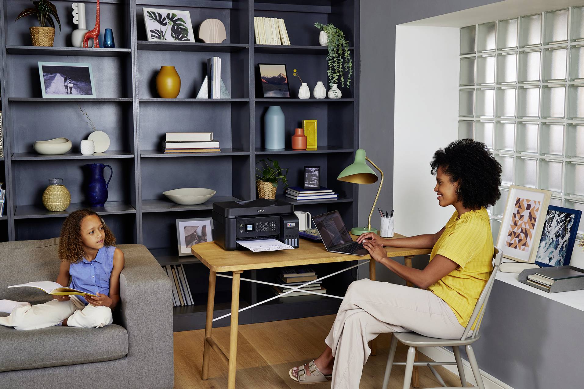 Eine Frau arbeitet an einem Laptop an einem Tisch mit einem Drucker, während ein Kind auf dem Sofa sitzt und ein Buch liest. Regal im Hintergrund.