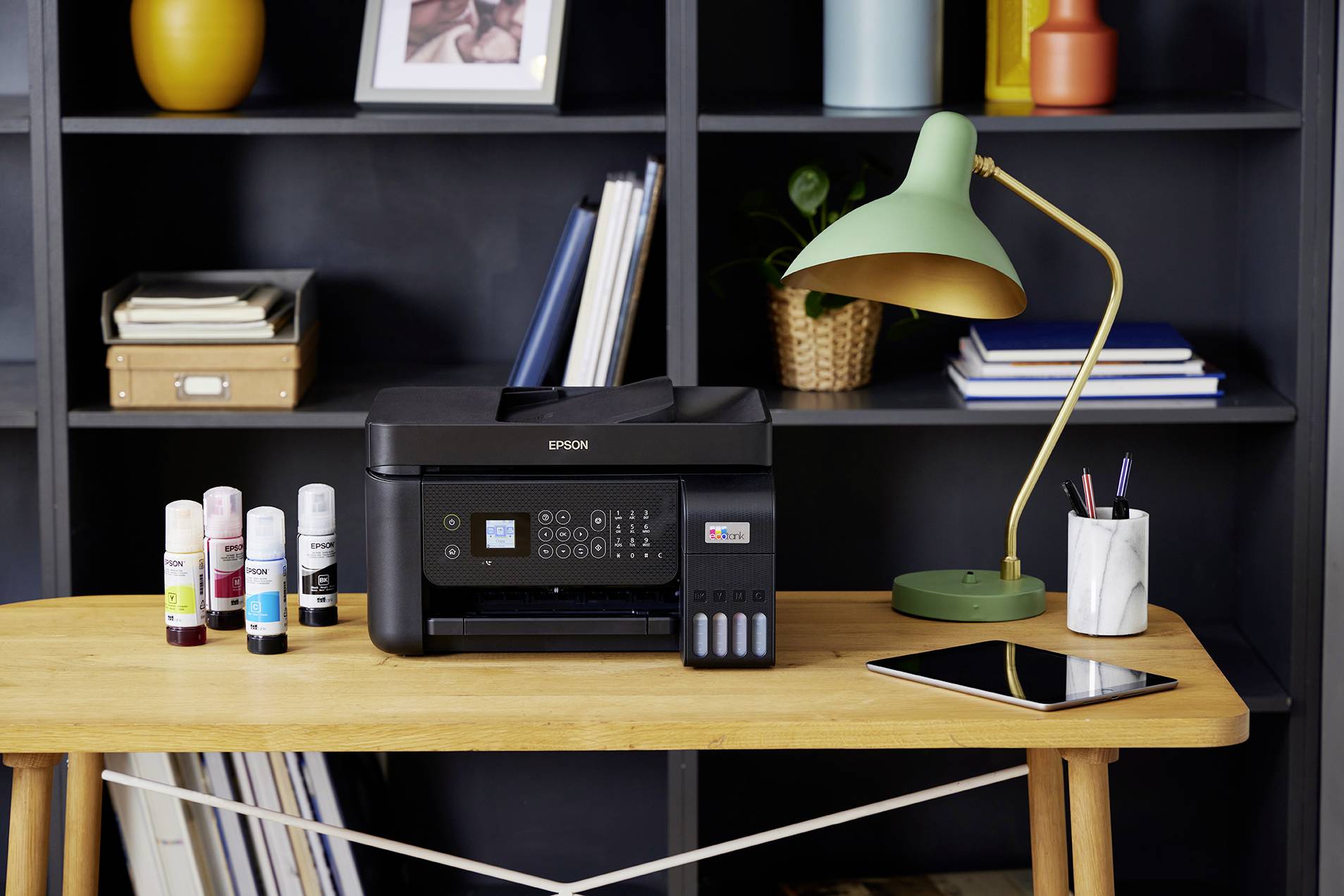 Ein Schreibtisch mit einem Multifunktionsdrucker, daneben Tintenflaschen. Im Hintergrund ein Bücherregal, auf dem Tisch eine Lampe und ein Tablet.