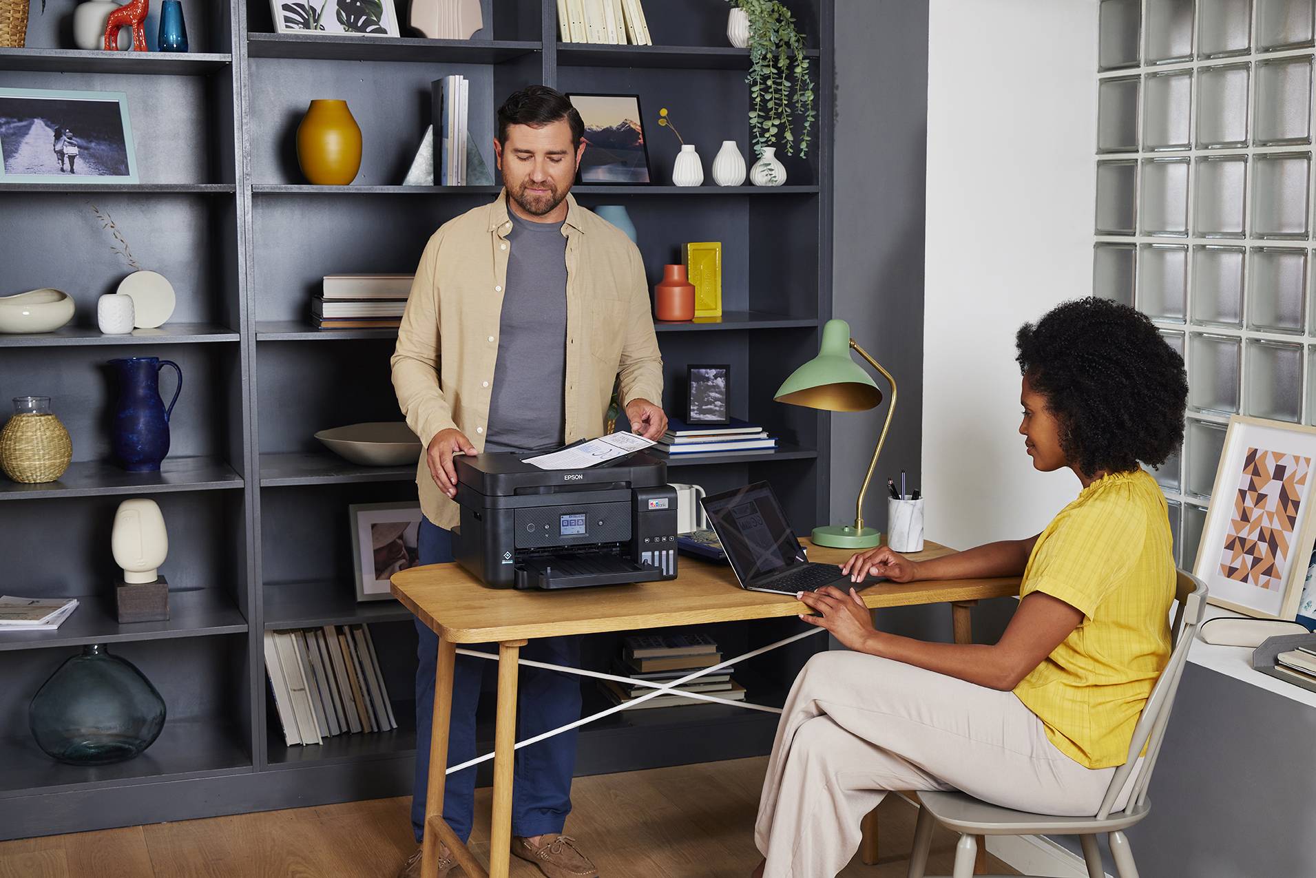 'Ein Mann bedient einen Drucker auf einem Holztisch, während eine Frau am Laptop arbeitet. Im Hintergrund ein großes Regal mit Dekor.'