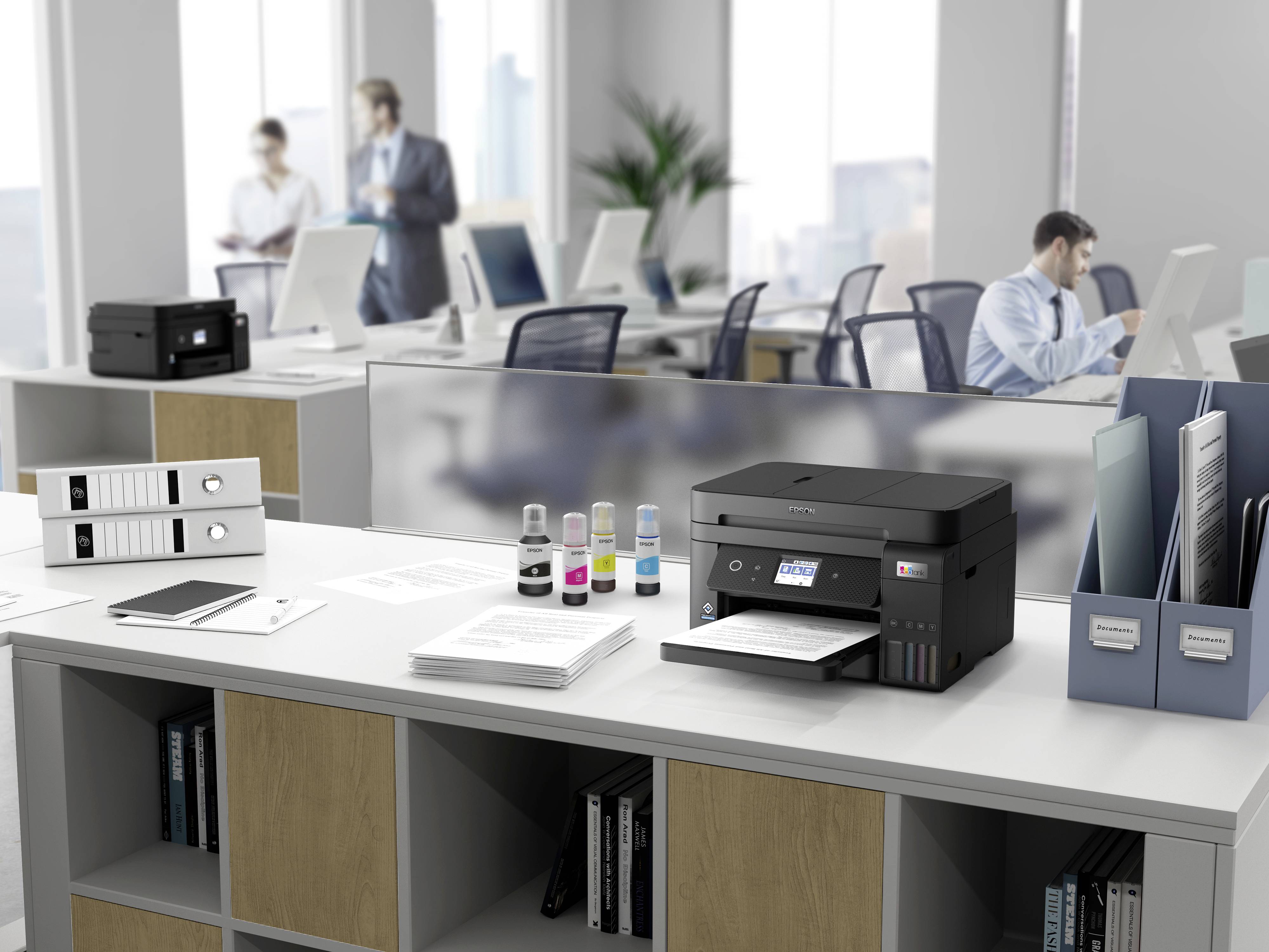 Drucker auf einem Schreibtisch im Büroumfeld mit Papieren und Tintenflaschen. Im Hintergrund arbeiten Personen an Computern.