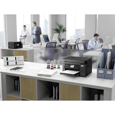 Drucker auf einem Schreibtisch im Büroumfeld mit Papieren und Tintenflaschen. Im Hintergrund arbeiten Personen an Computern.