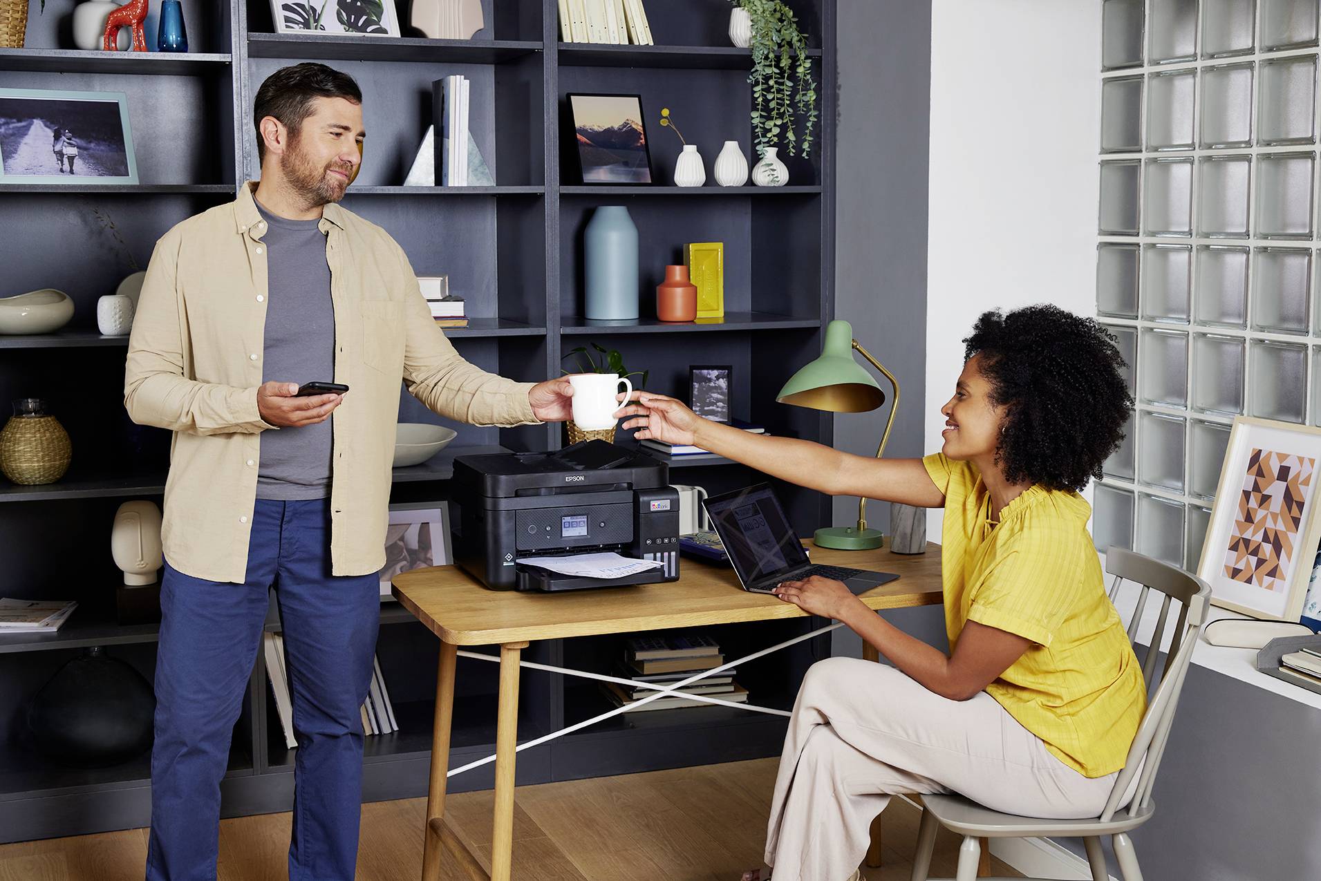 Ein Mann reicht einer Frau eine Tasse in einem Büro mit Bücherregal. Die Frau sitzt mit einem Laptop, der Mann steht mit einem Smartphone.