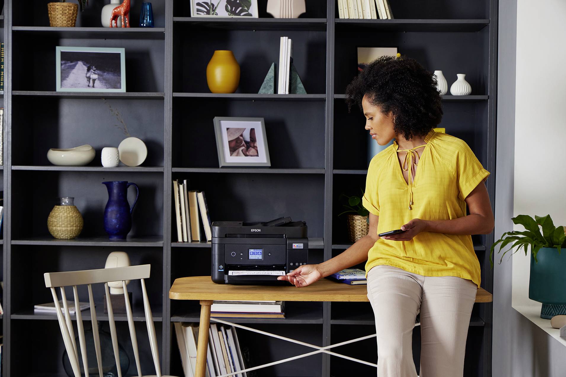 Eine Frau in gelber Bluse bedient einen Drucker auf einem Tisch vor einem Regal voller Dekorobjekte. Sie hält ein Smartphone in der Hand.