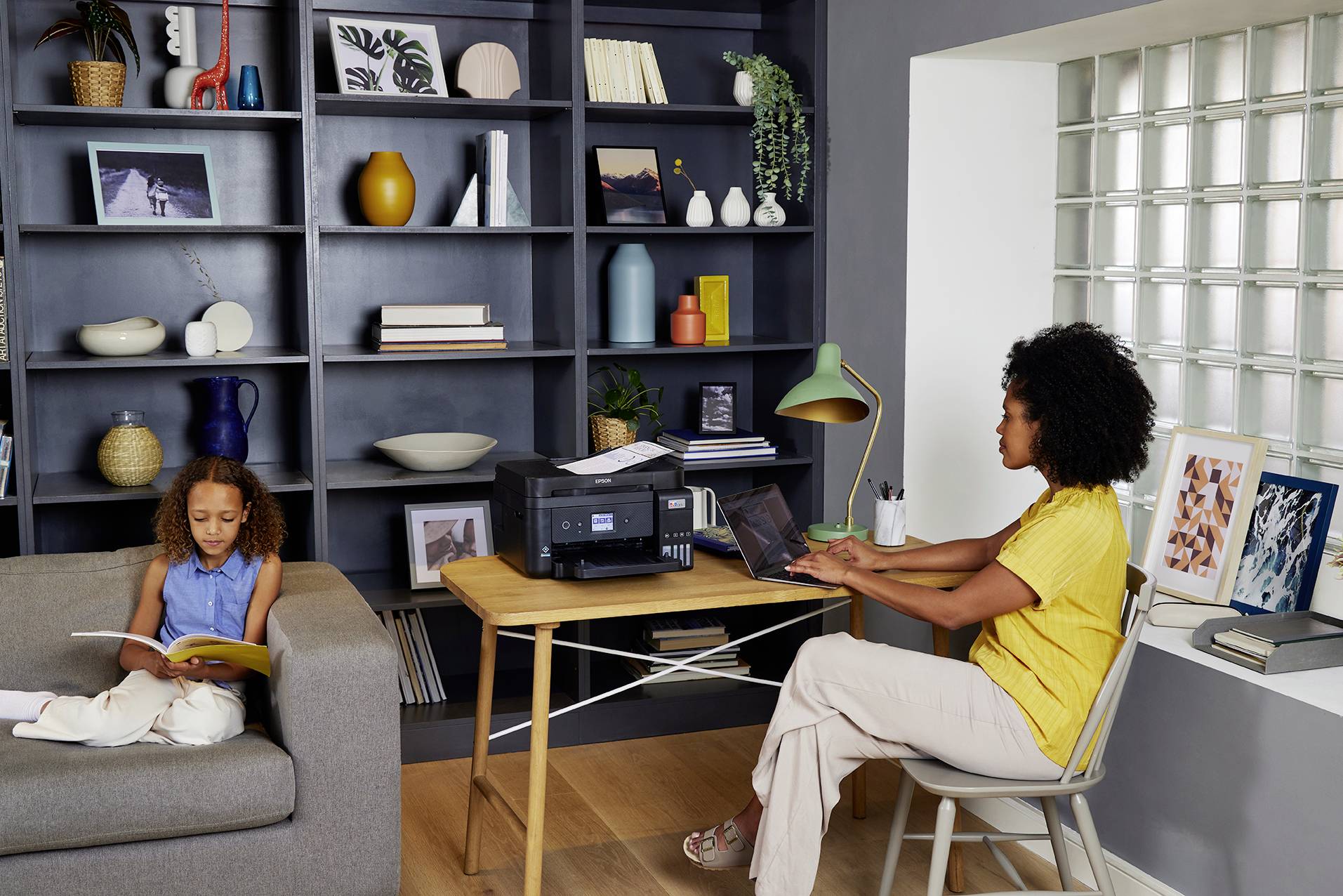 Eine Frau arbeitet an einem Laptop am Schreibtisch neben einem Drucker. Ein Kind sitzt auf dem Sofa und liest ein Buch. Regal im Hintergrund.