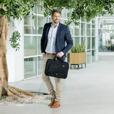 Ein Mann in einem Anzug steht lächelnd neben einem Baum in einem modernen Gebäude, hält eine schwarze Laptoptasche in der Hand.