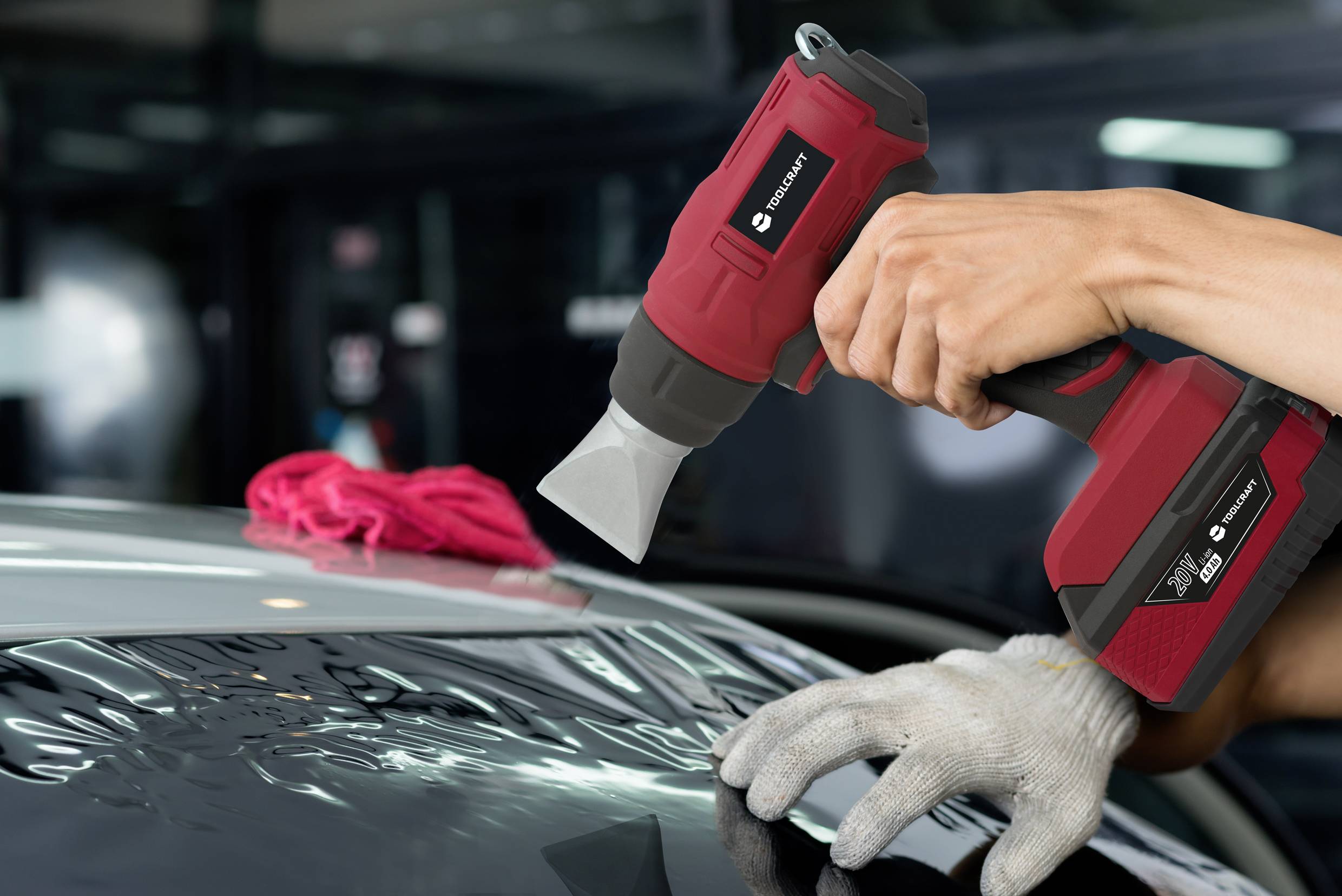 Eine Person hält ein rotes Heißluftgebläse und bearbeitet ein Autofenster. Ein rotes Tuch liegt auf dem Dach des Autos.