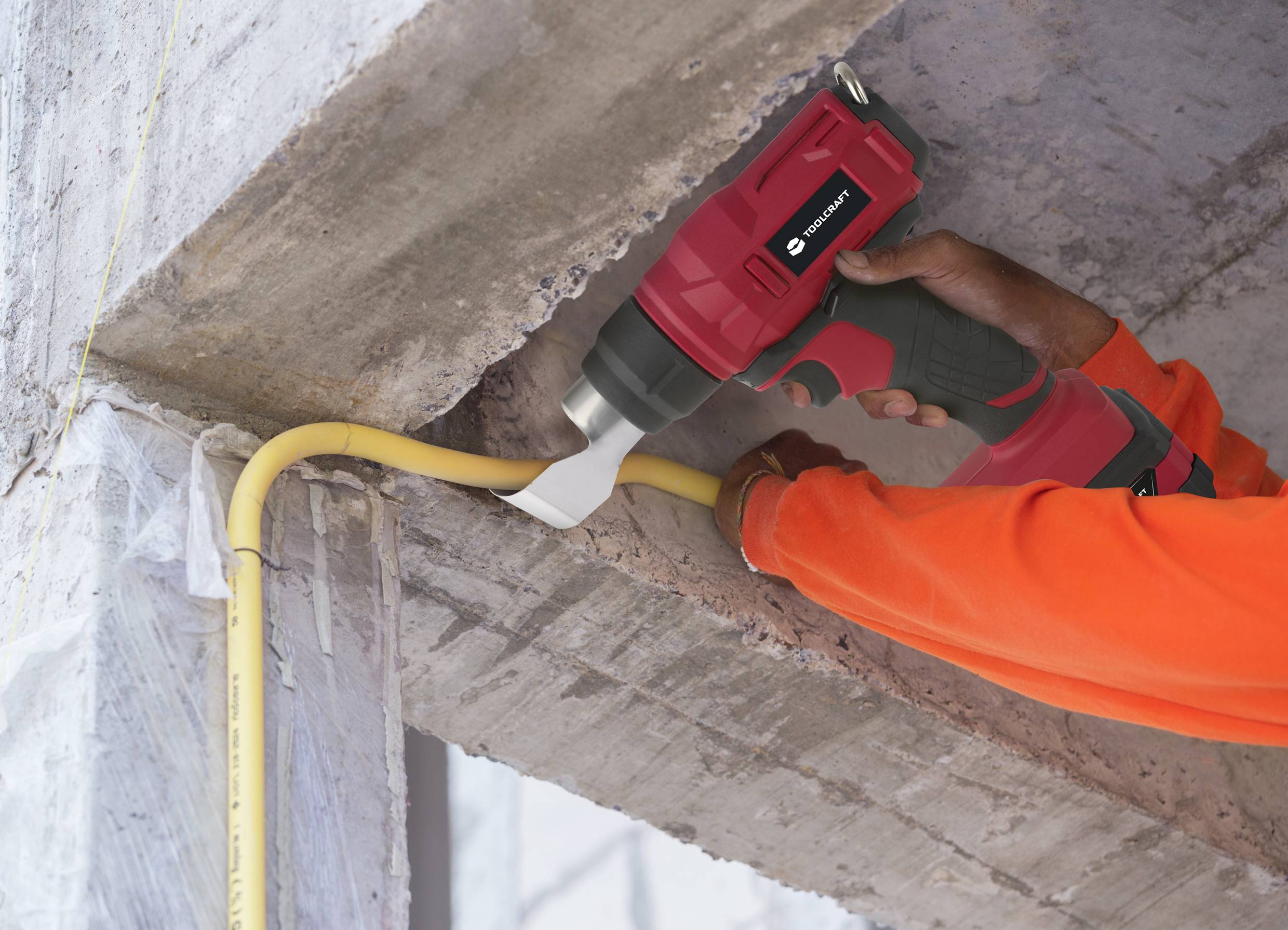 Person in orange Arbeitsoverall benutzt elektrischen Abisolierer, um gelbes Kabel an Betonwand zu bearbeiten.