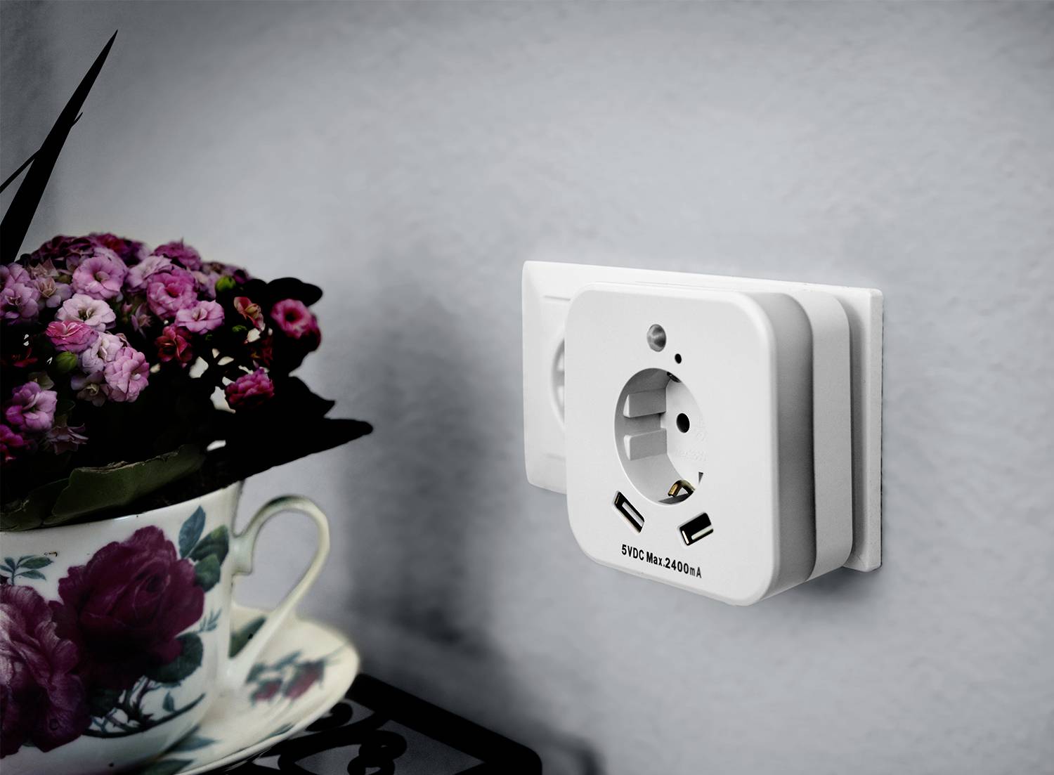 Weiße Steckdose mit zwei USB-Anschlüssen an einer grauen Wand; daneben steht eine Tasse mit Blumenmuster und rosa Blüten.